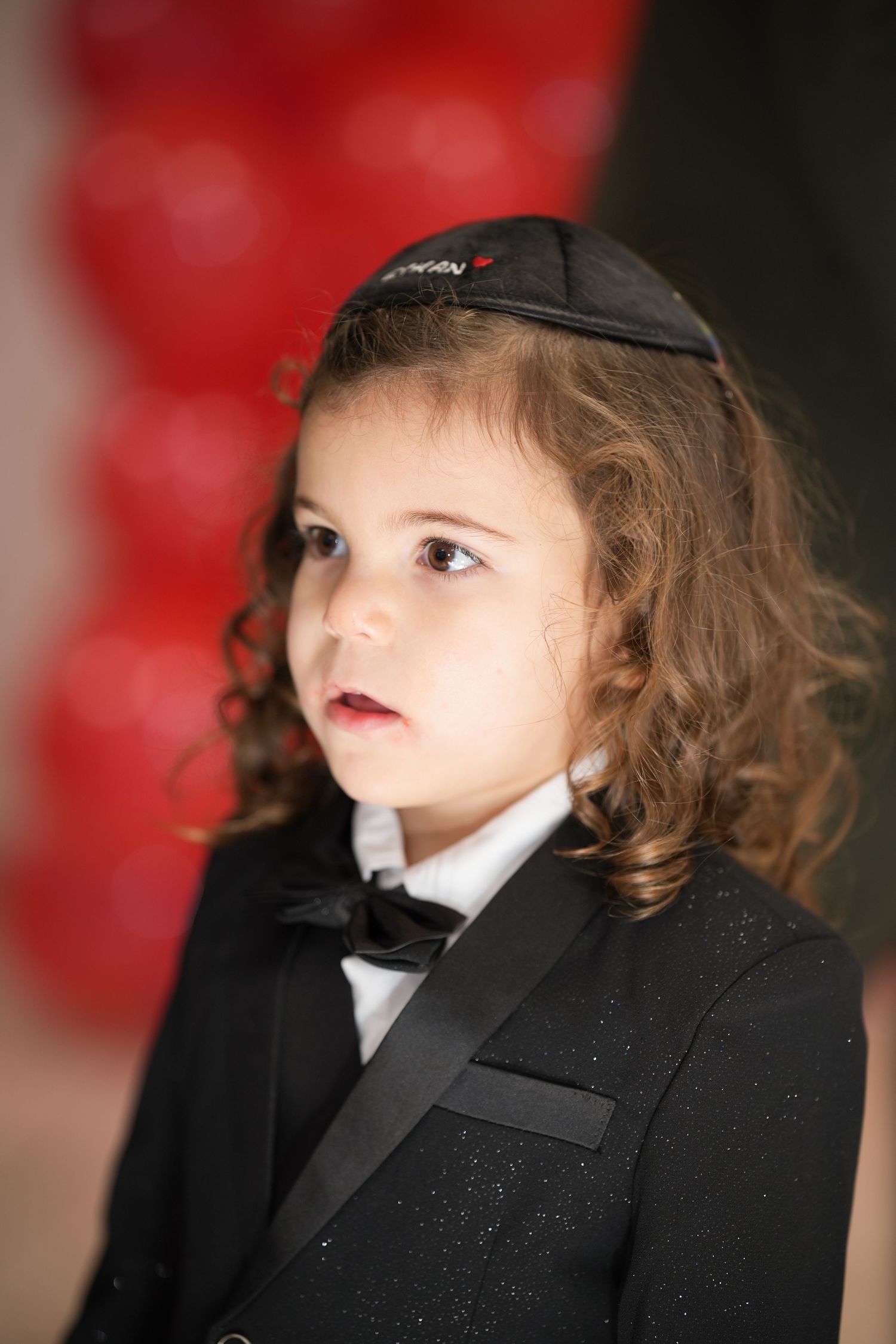 Adorable young child with curly hair wearing black tuxedo and kippah at a party.