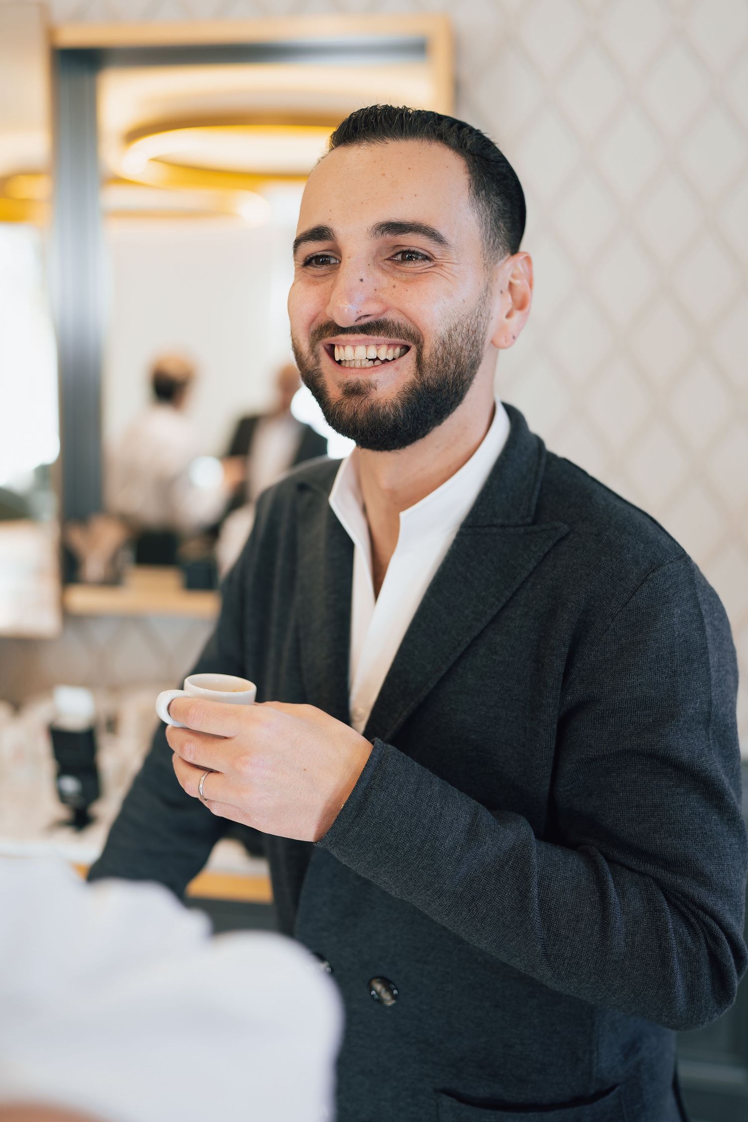 Smiling bearded man in dark blazer holding espresso cup in a bright modern cafe setting.