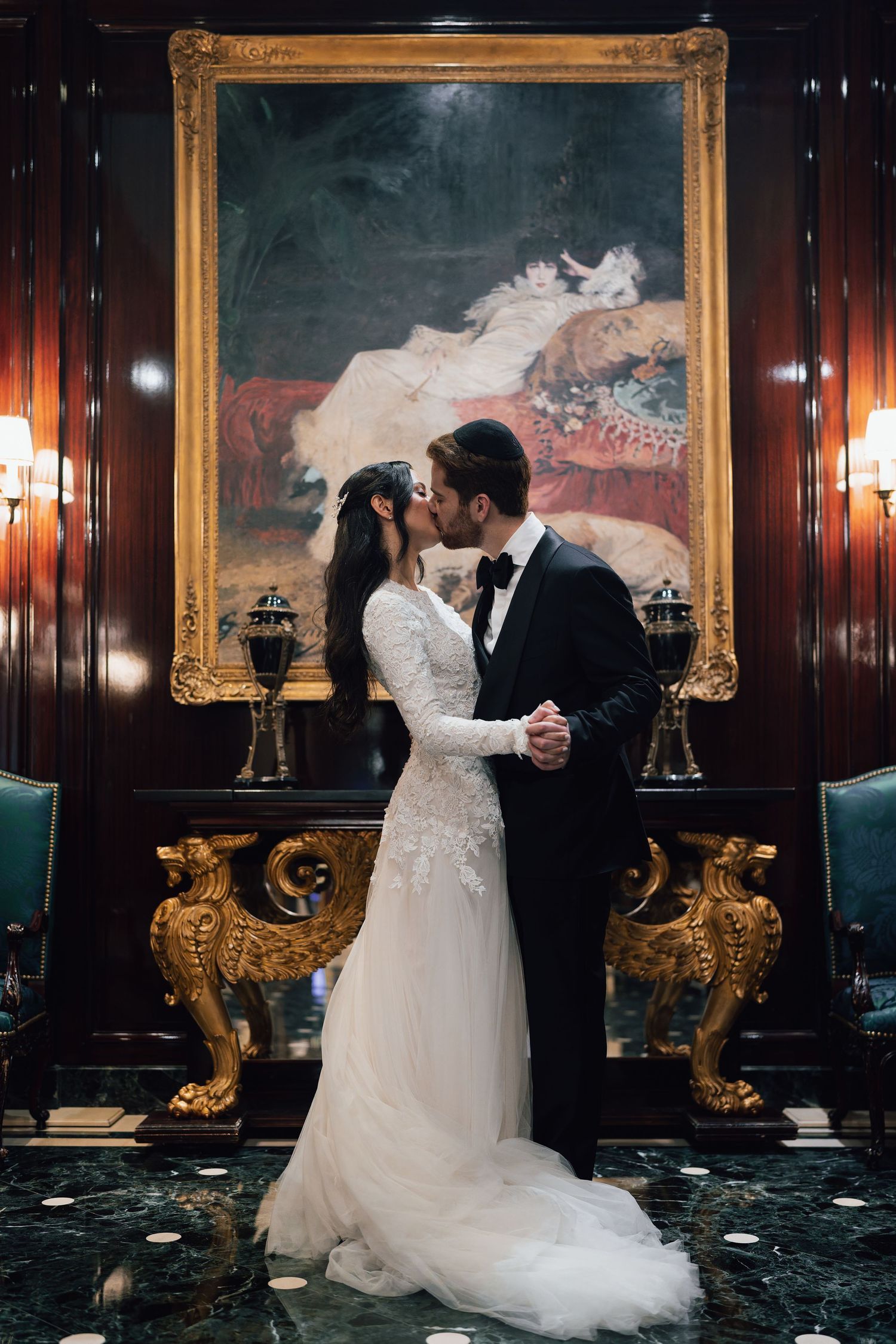 Bride and groom sharing a romantic kiss in an opulent room with golden decor and classical oil painting backdrop.
