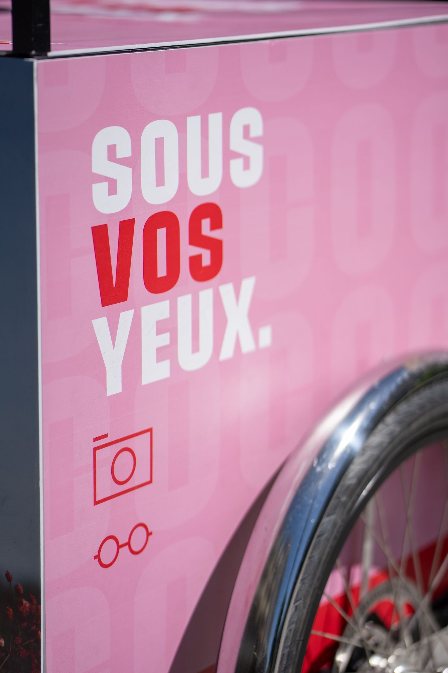 Pink sign with French text 'Sous Vos Yeux' featuring camera and glasses icons, beside a chrome bicycle wheel.