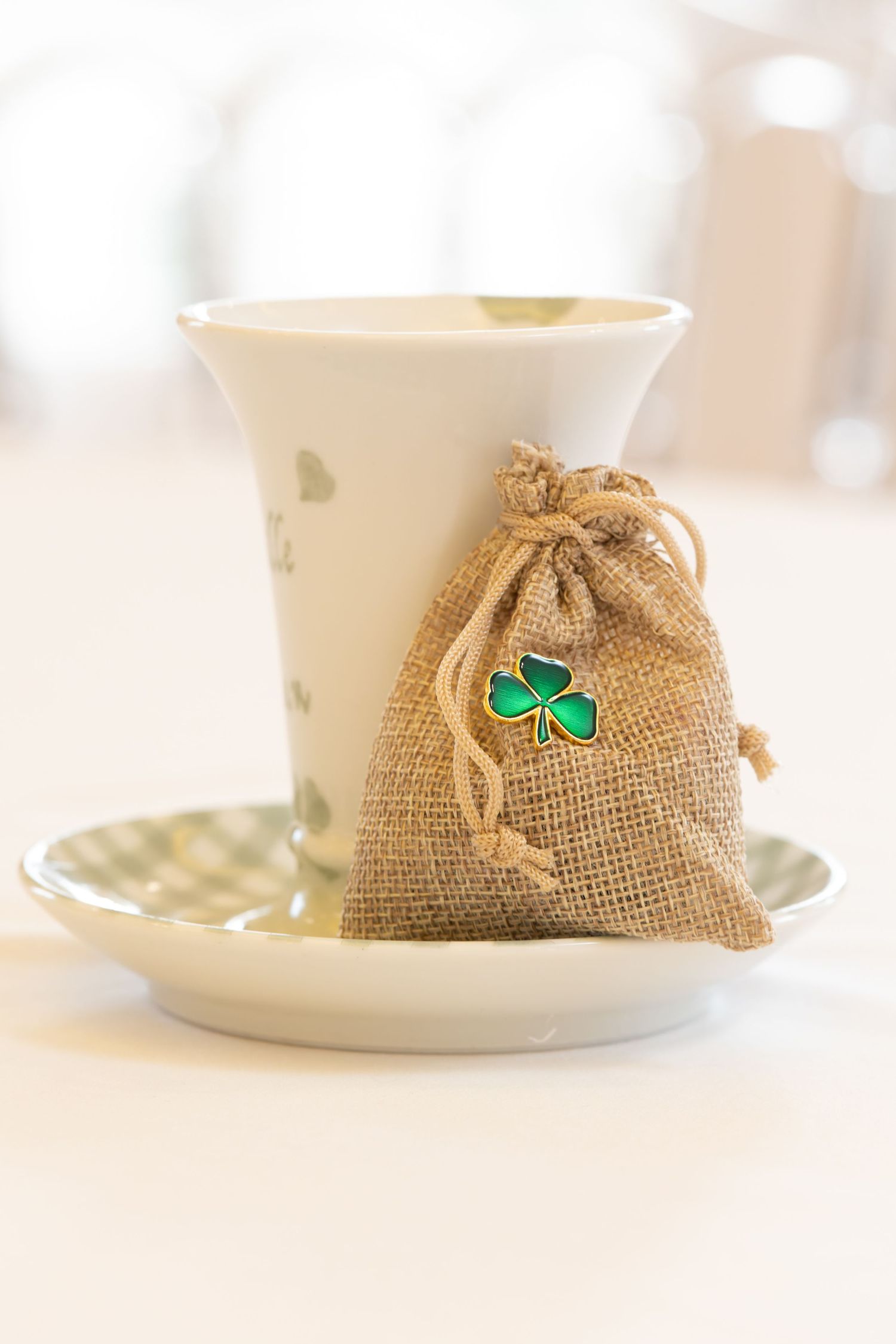Burlap gift bag with green shamrock charm leaning against white ceramic cup on decorative saucer.