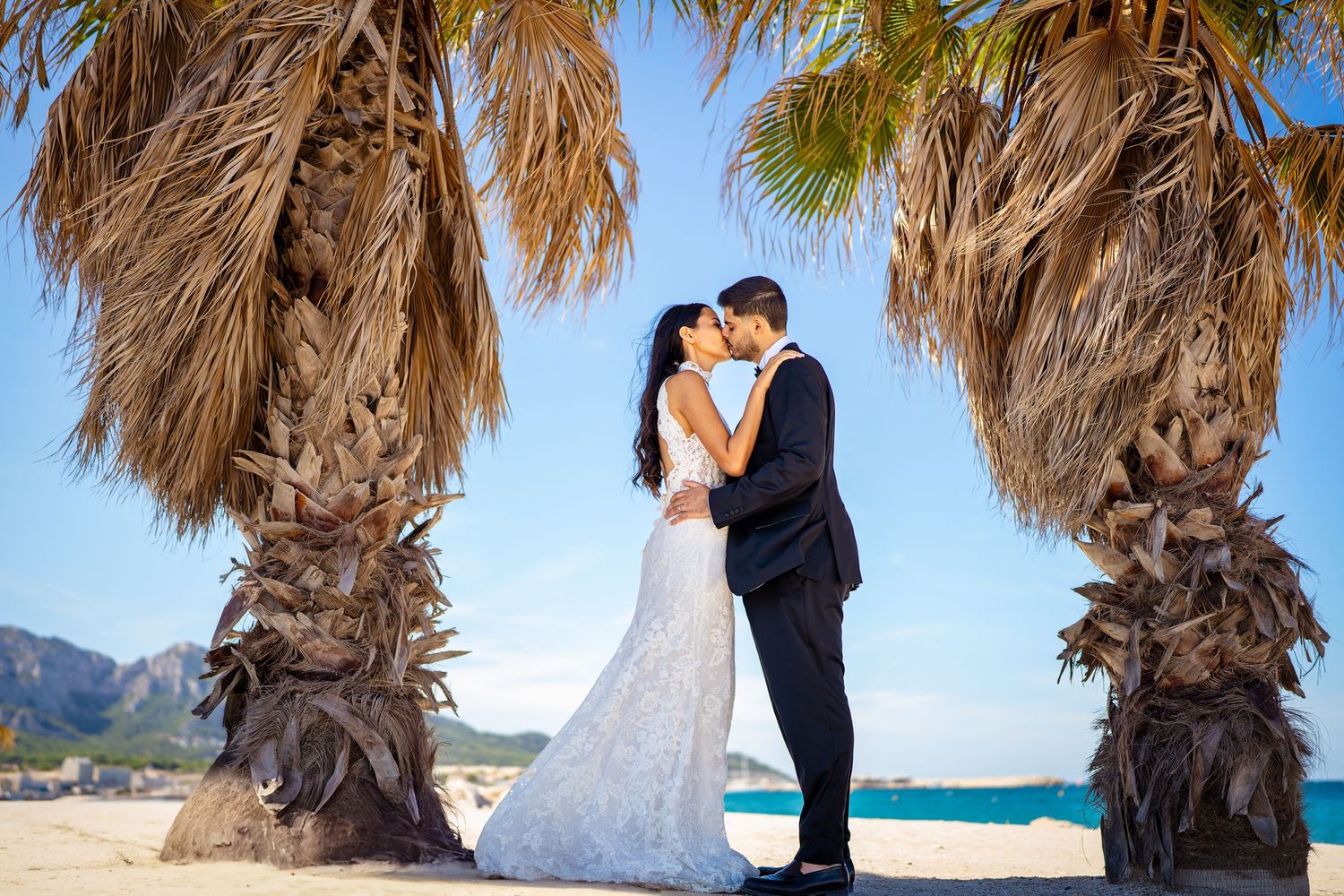 Wedding couple sharing a romantic kiss beneath tall palm trees on a sunny Mediterranean beach.