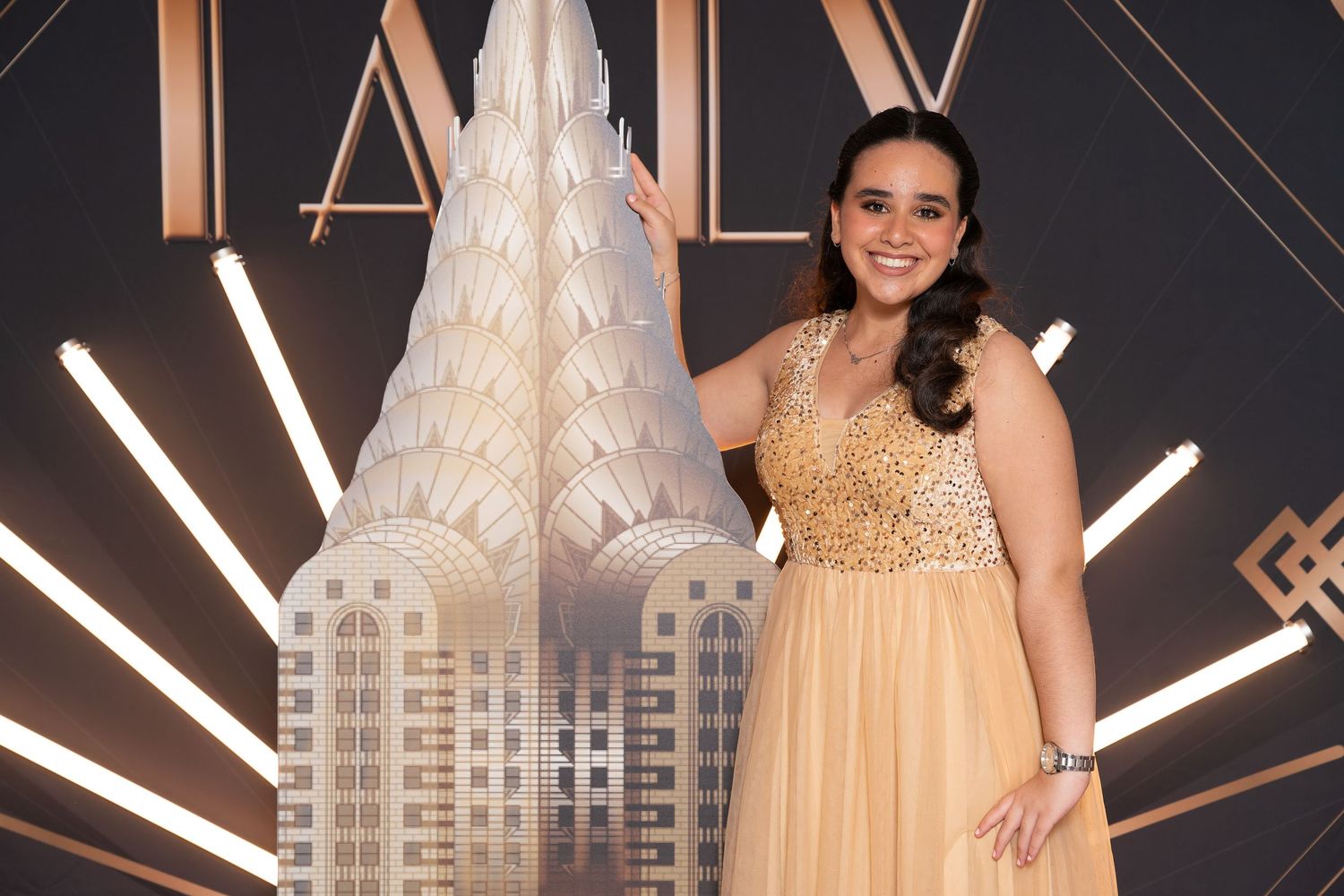 Woman in gold beaded gown posing in front of Art Deco Chrysler Building backdrop at formal event.