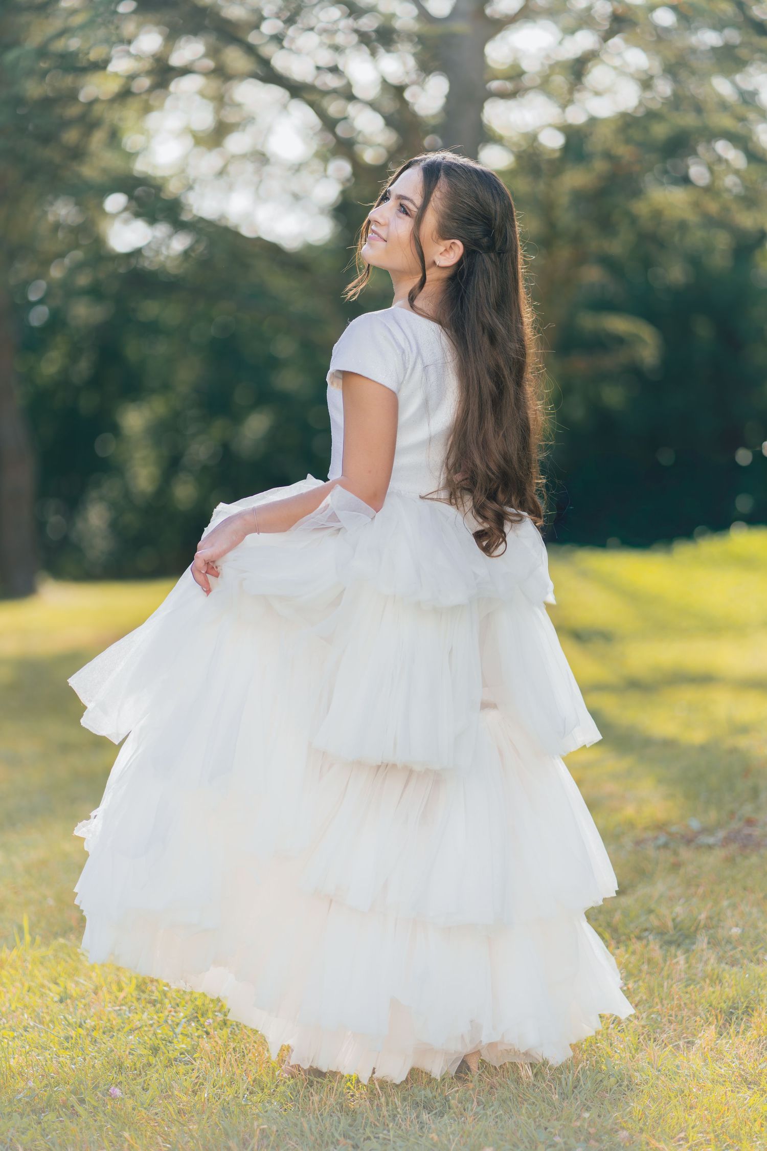 Young girl in white tiered tulle gown twirling in sunlit green park setting.