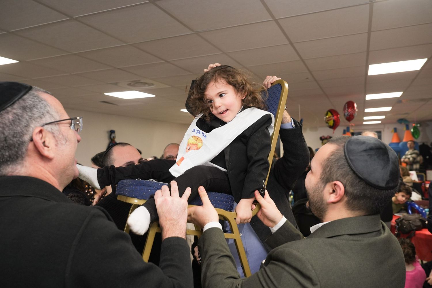 Jewish child being lifted on a chair during a traditional celebration ceremony with adults.