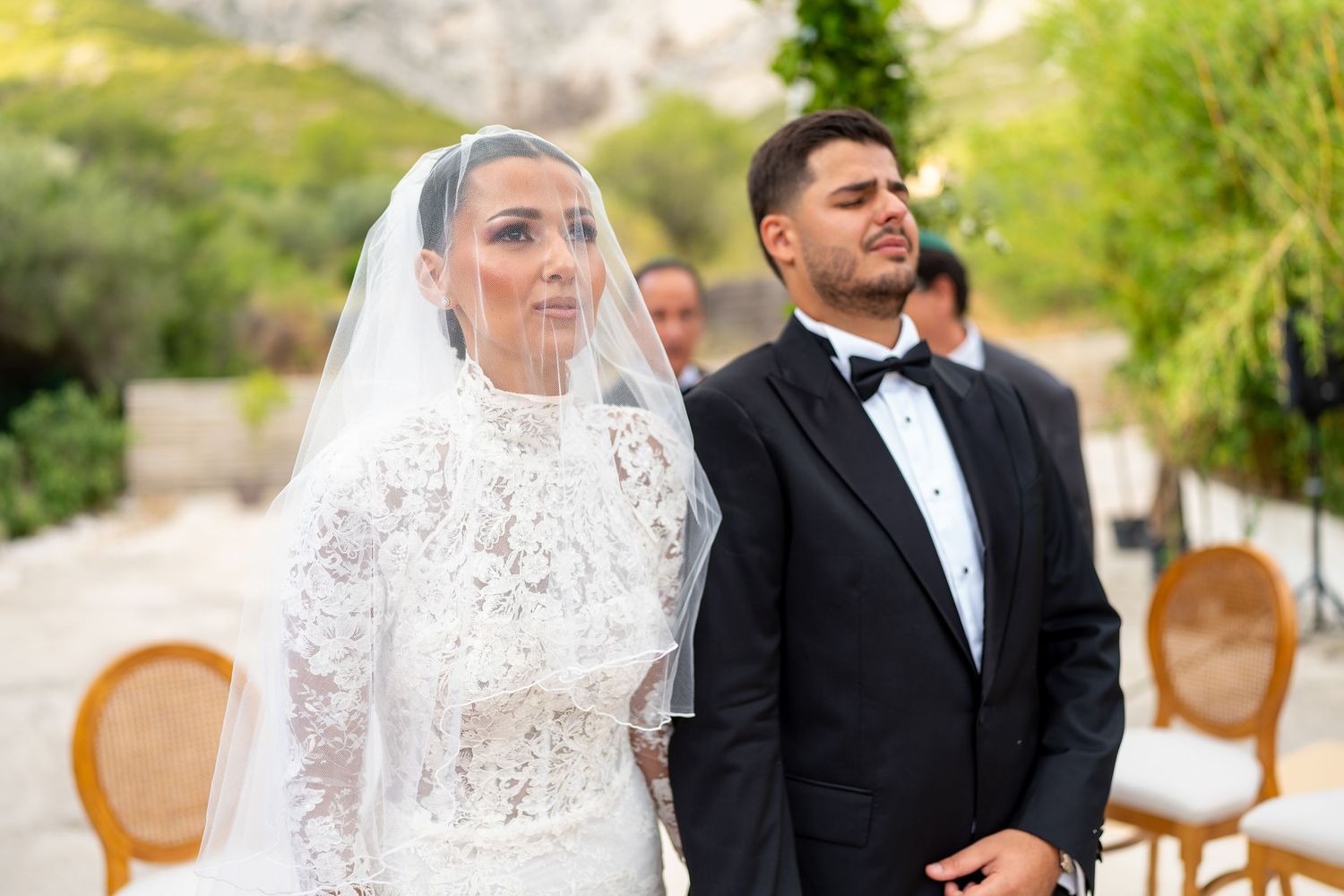 Bride in intricate lace veil and groom in tuxedo standing at an elegant outdoor wedding ceremony.