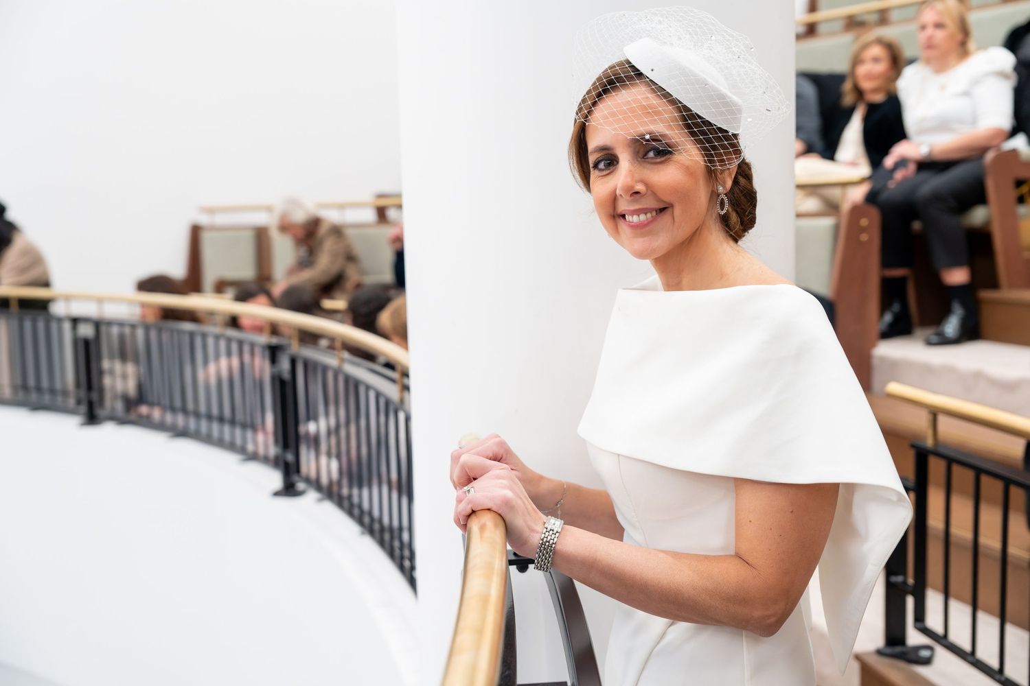 Elegant woman in white bridal gown with cape detail and veil stands on curved staircase railing indoors.