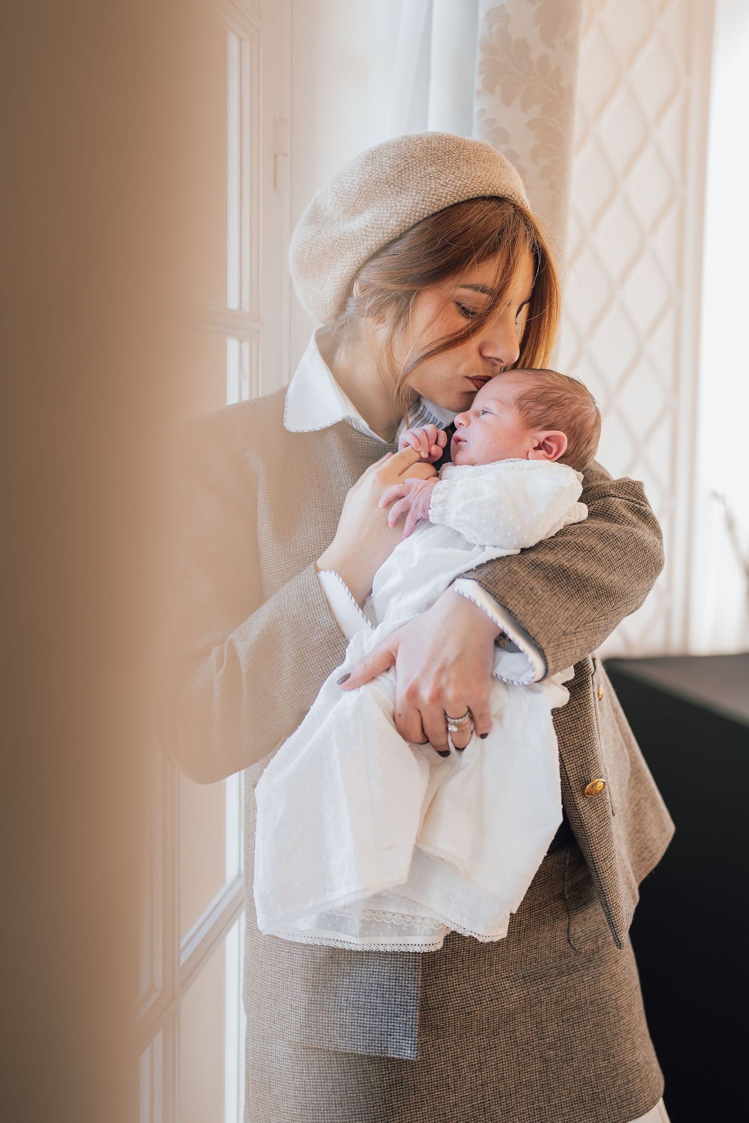A woman in a beige beret and tweed coat tenderly kisses her newborn baby dressed in white near a bright window.