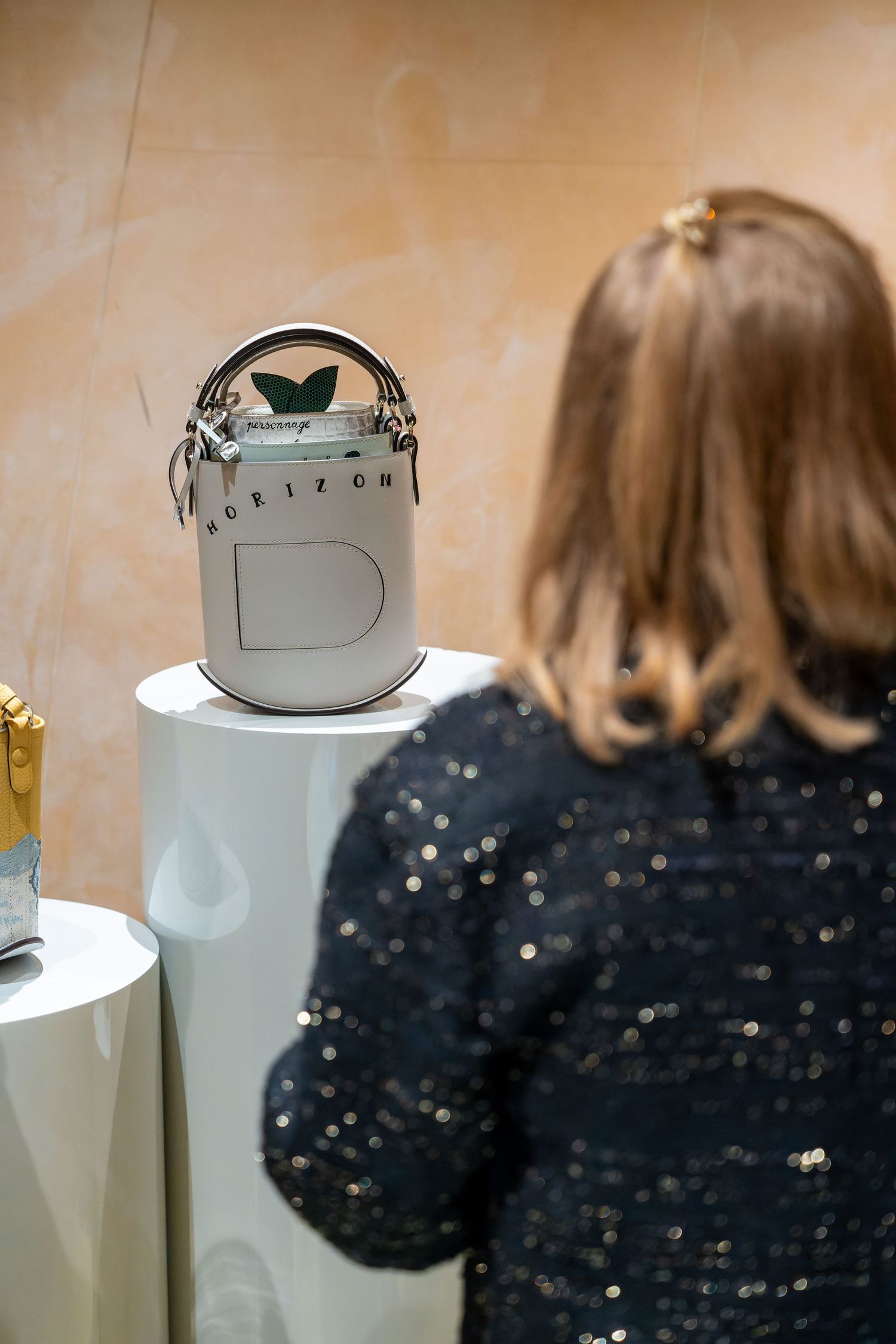 Woman in sequined jacket admiring a white Delvaux Horizon bucket bag displayed on a white pedestal.