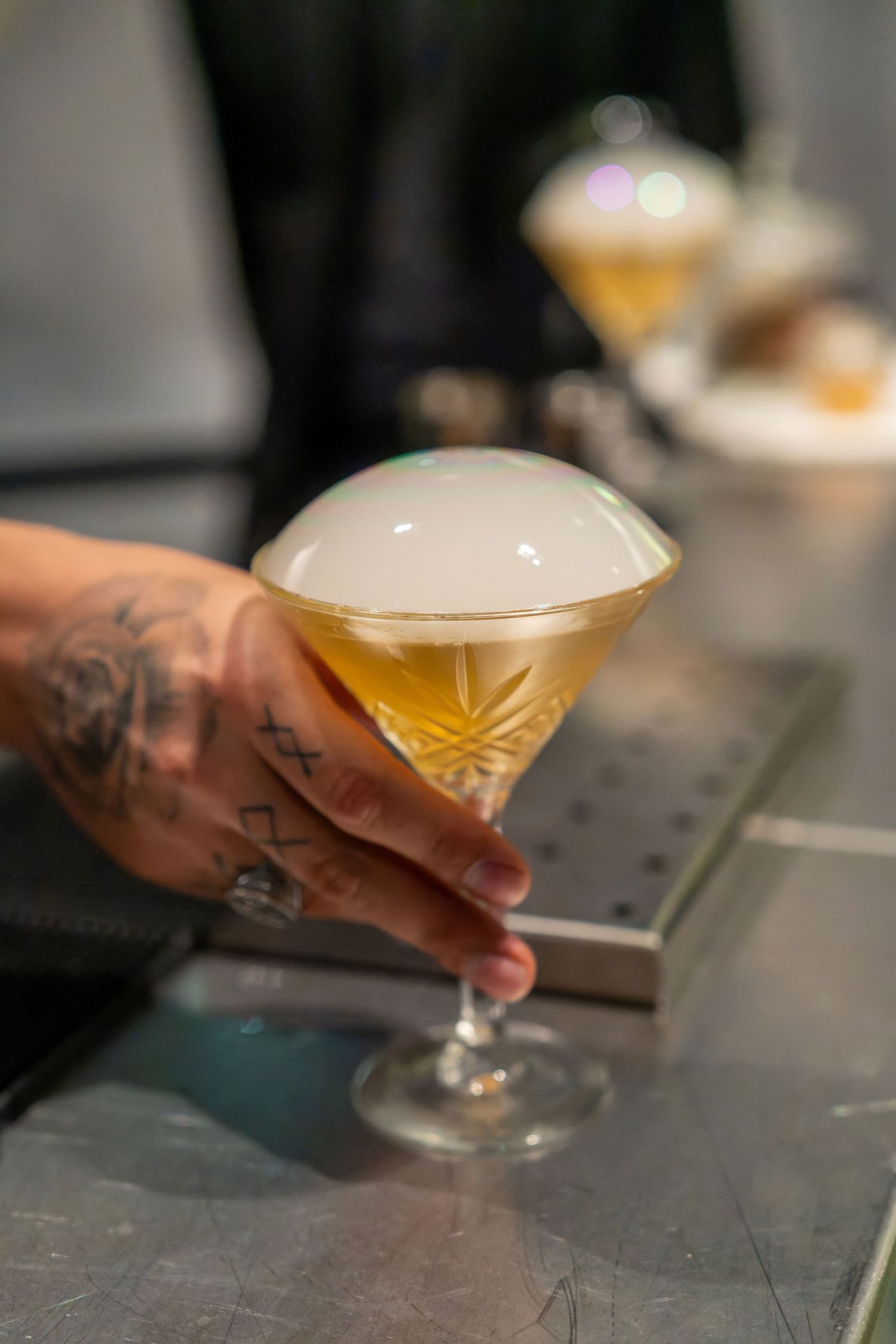Tattooed hand holding a crystal coupe glass with amber cocktail topped with white foam at a bar.