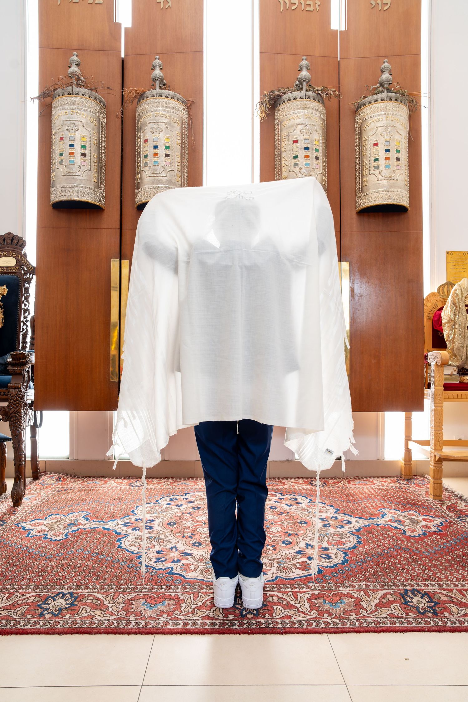 Person wrapped in a white Jewish prayer shawl tallit standing before Torah scrolls in a synagogue.
