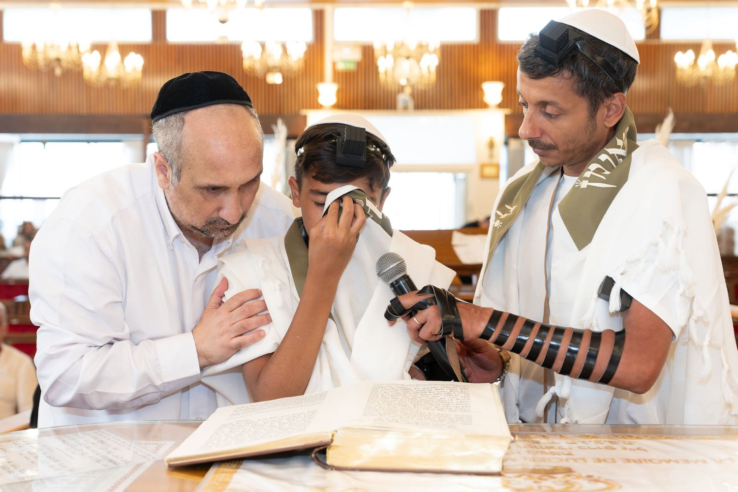 Boy reads Torah at Bar Mitzvah flanked by rabbi and father in synagogue.