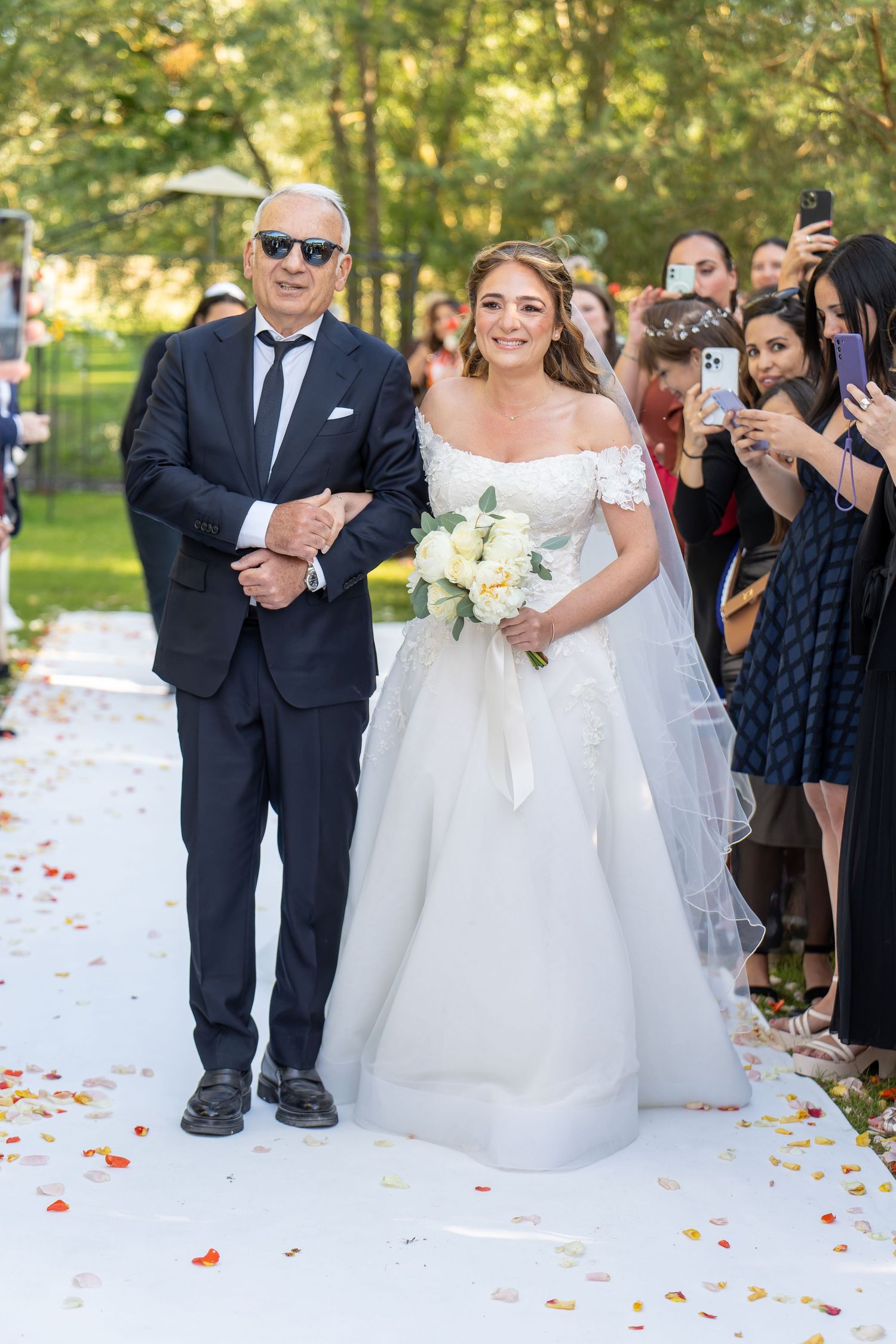 Bride in elegant off-shoulder white gown walks down aisle with father during outdoor wedding ceremony.