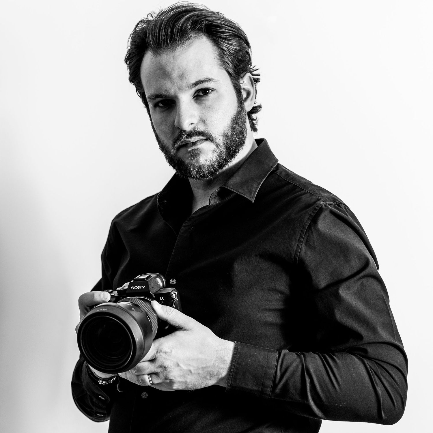 Professional male wedding photographer in black shirt holding a Sony camera, captured in black and white portrait.