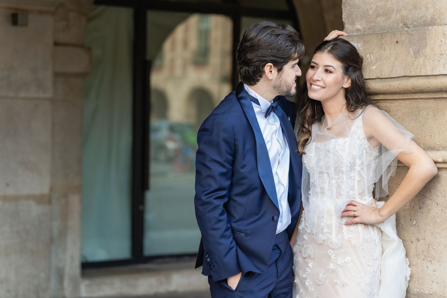 Elegant wedding couple posing by stone columns, groom in navy suit, bride in white lace gown, sharing a romantic moment.