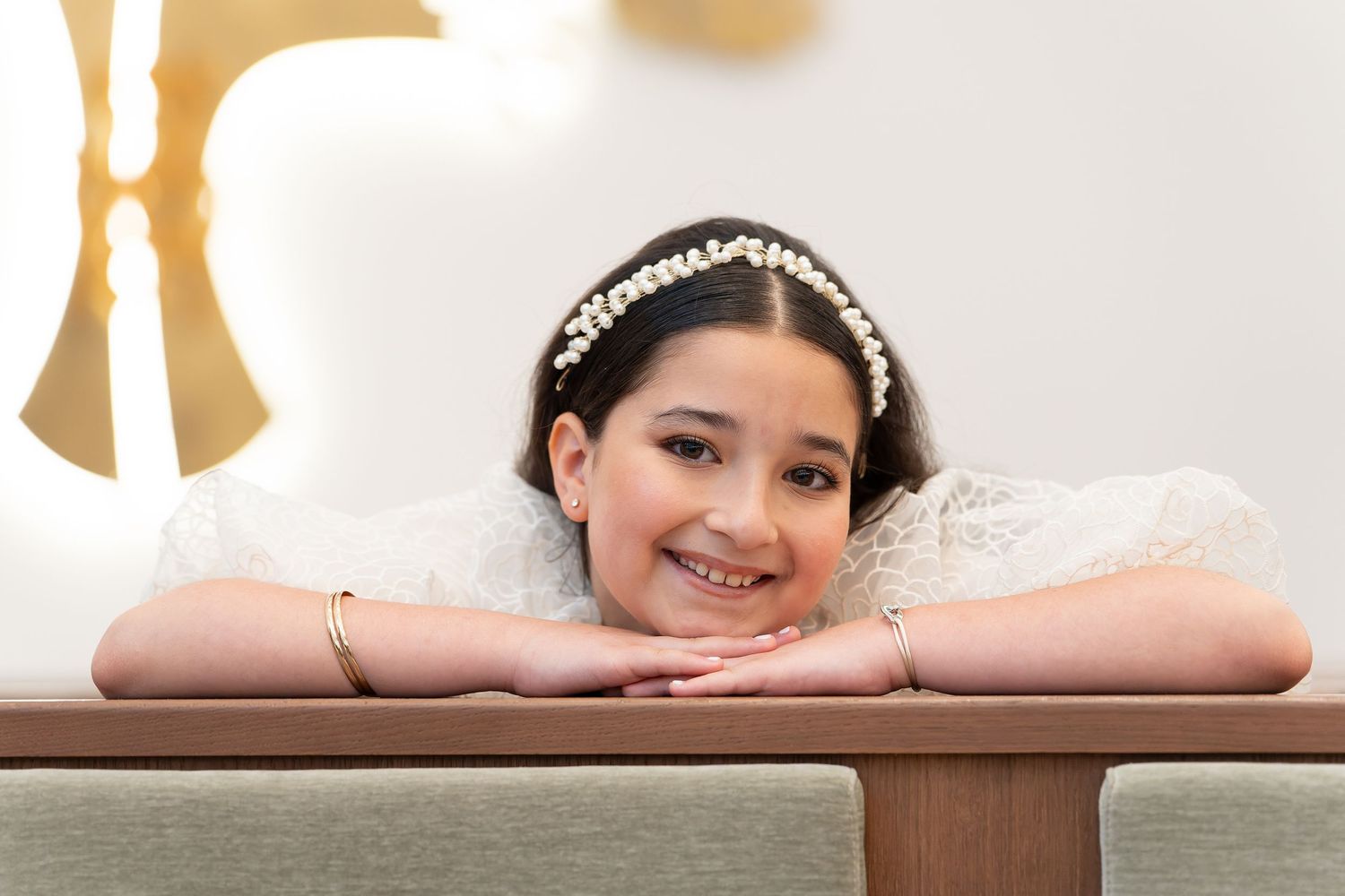 Smiling young girl in white dress with pearl headband resting chin on arms at bench.