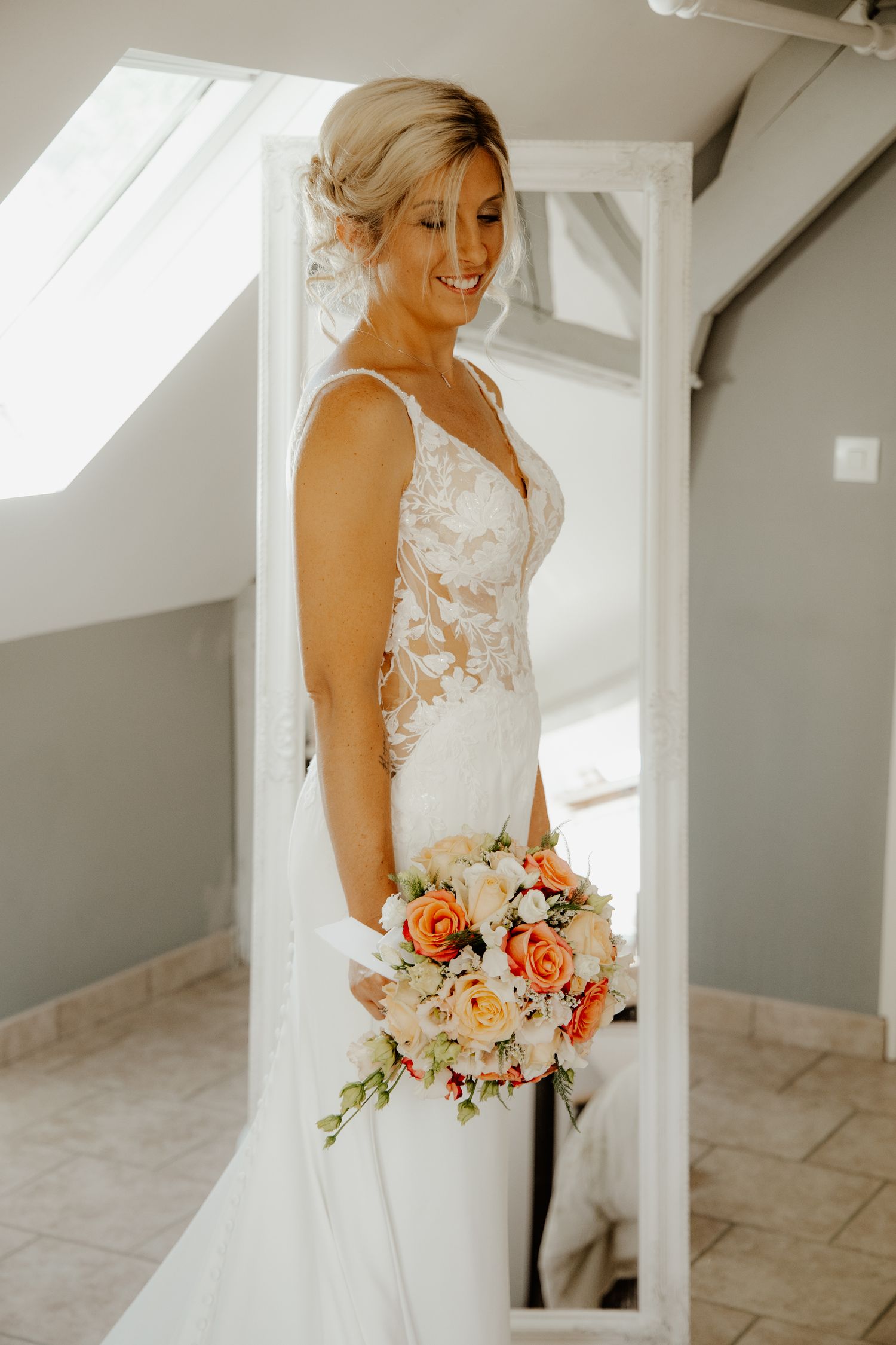 Portrait lumineux de la mariée souriante en robe blanche près d'une fenêtre durant les préparatifs.