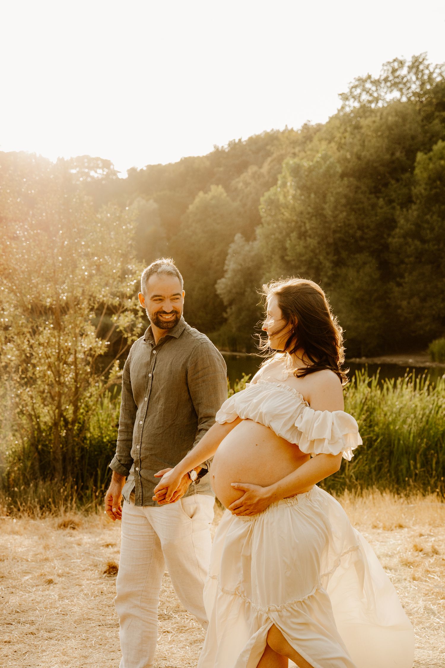 Couple se promenant au bord d’un étang en se regardant dans les yeux, la femme enceinte tenant son ventre.
