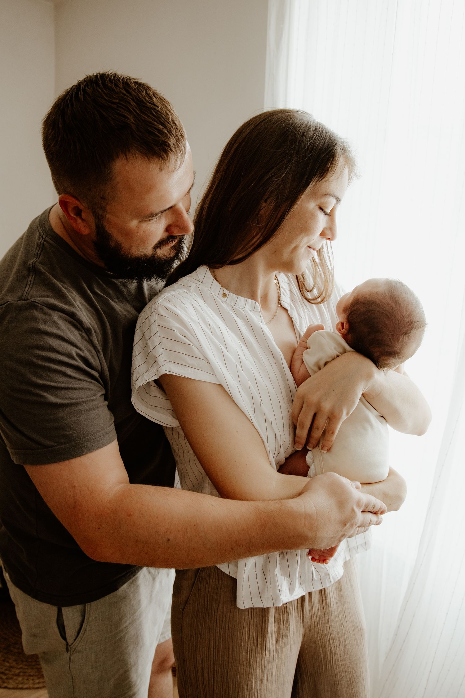 Un jeune couple de parents se serre tendrement dans les bras en regardant leur bébé.