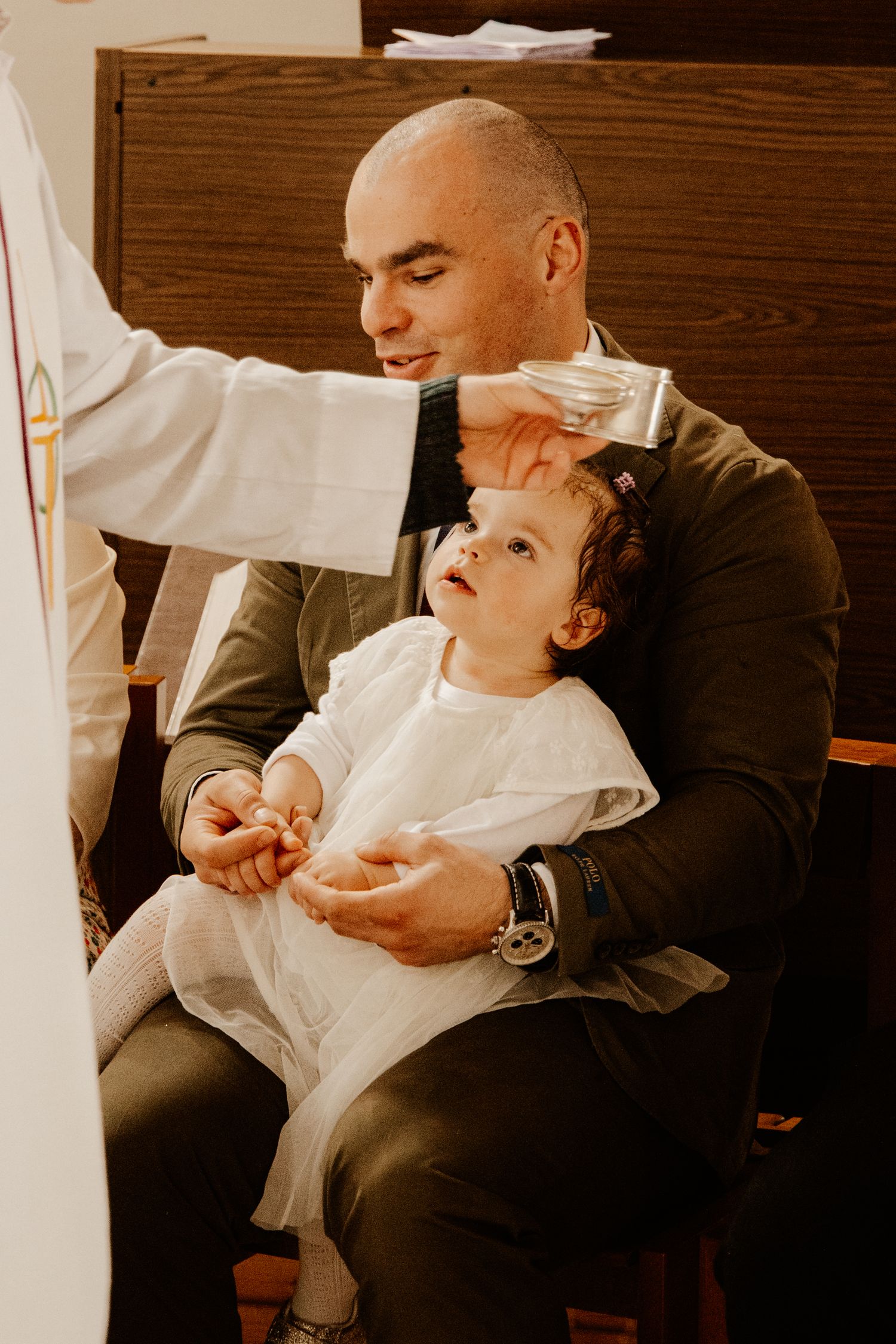 petit fille assise sur les jambe de son père et qui se fait baptiser par le prêtre