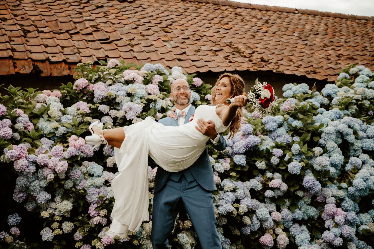 Le marié porte sa femme dans les bras en rigolant devant un mur de fleurs en briques. Séance photo de mariage.