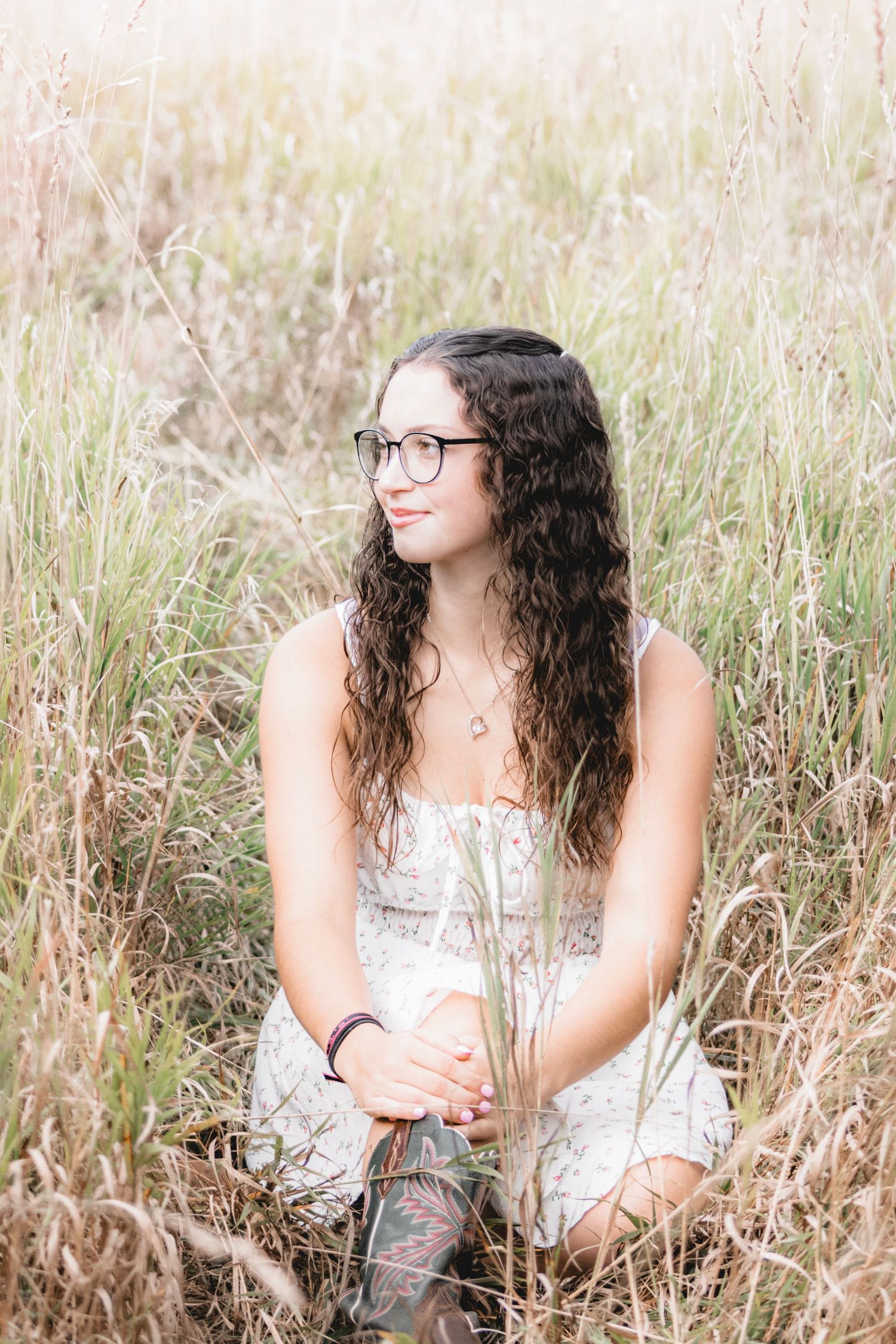 Young woman with curly hair and glasses sits in tall golden grass field wearing a white floral dress, smiling softly.
