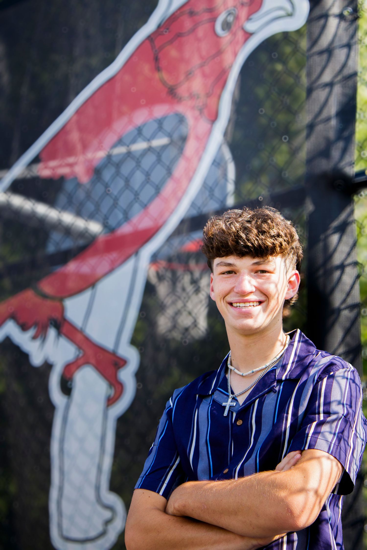 Smiling teen boy with curly hair crosses arms in front of a colorful cardinal mascot mural on a chain-link fence.