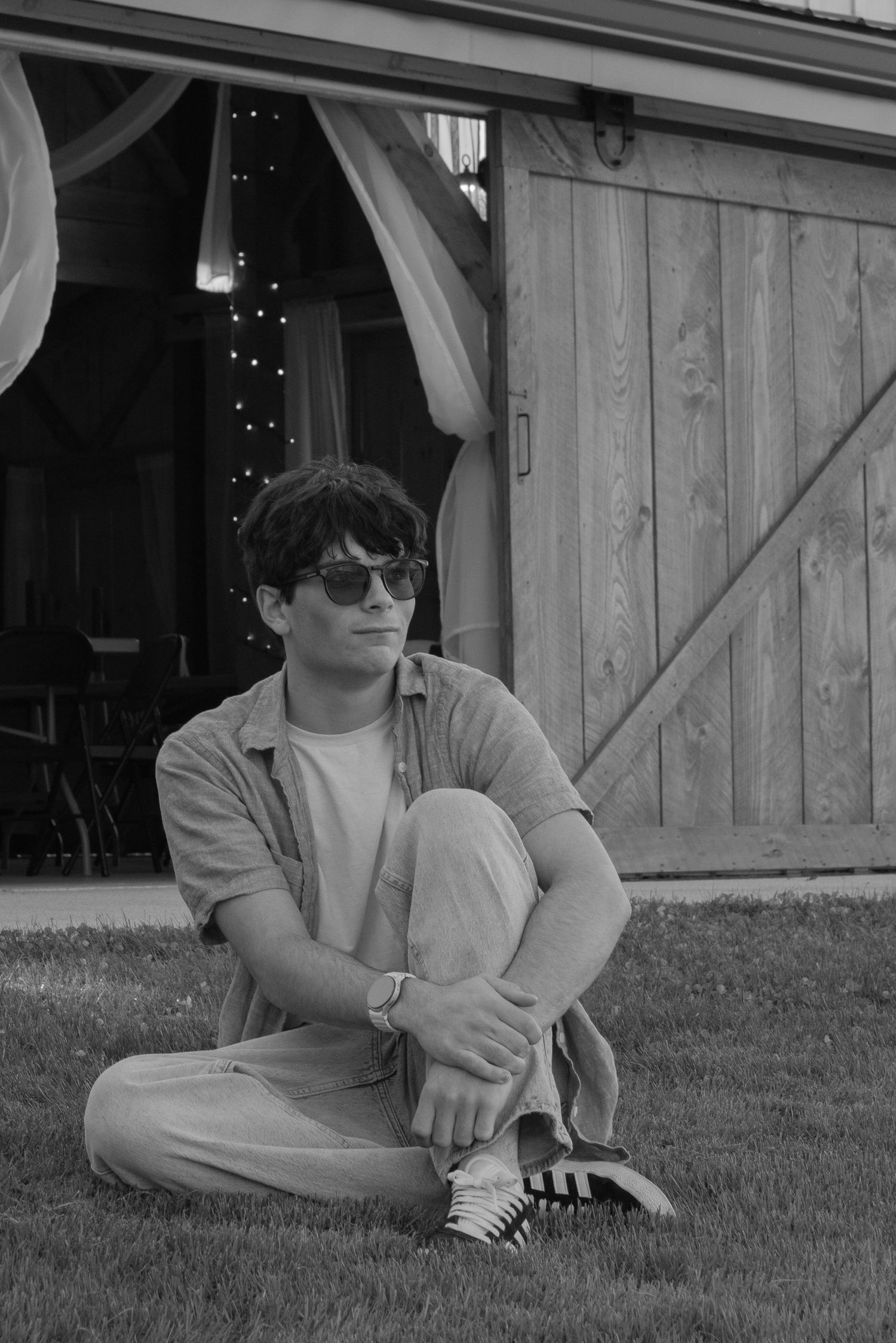 Young man in sunglasses sitting cross-legged on grass in front of a rustic barn in black and white.
