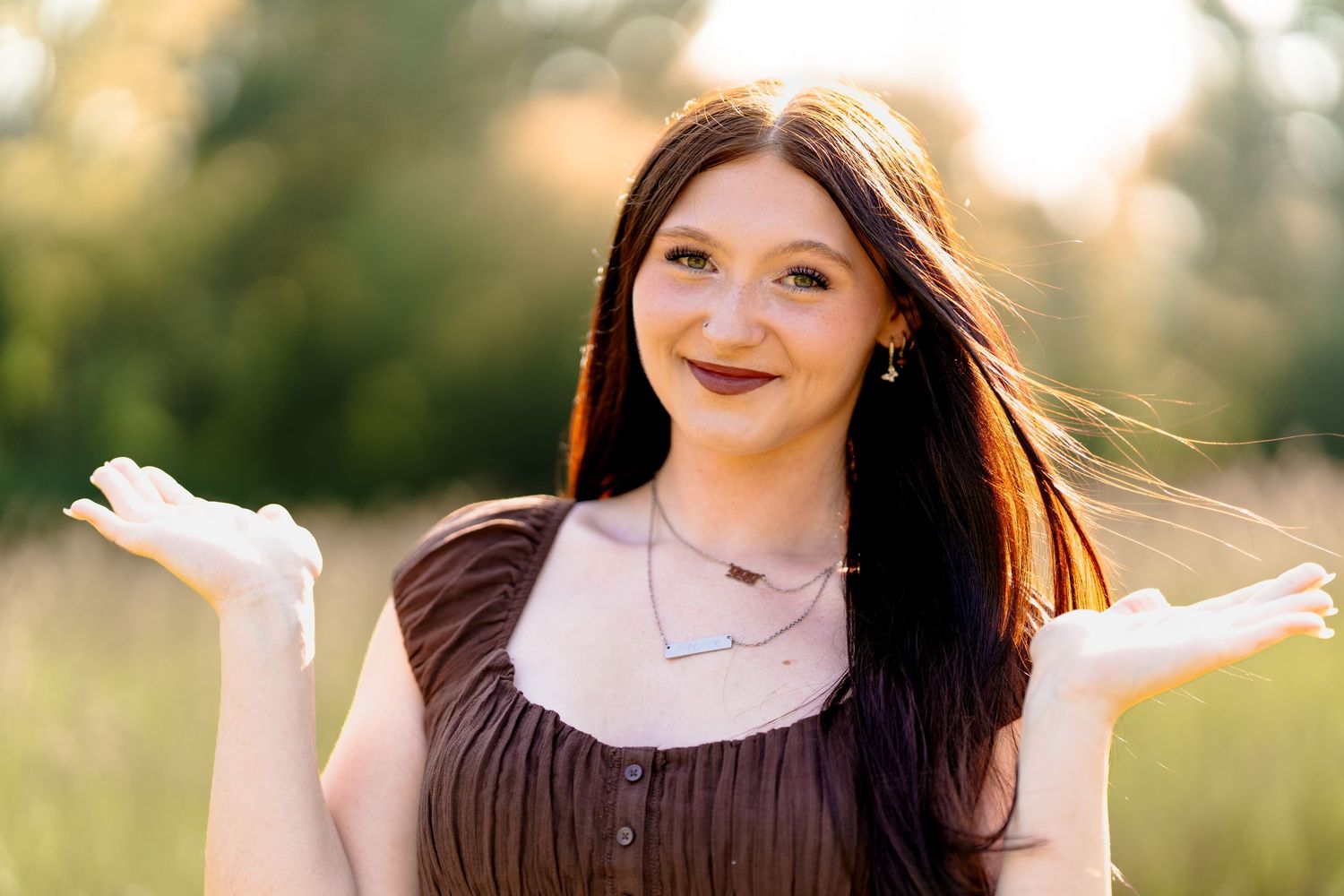 Young woman with long dark hair shrugging outdoors in golden hour light, wearing brown dress and layered necklaces.