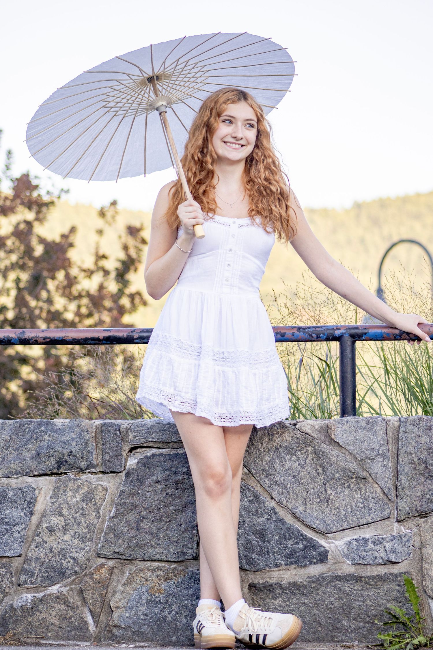 Young woman with curly red hair in white dress holding a parasol, leaning on stone wall outdoors.