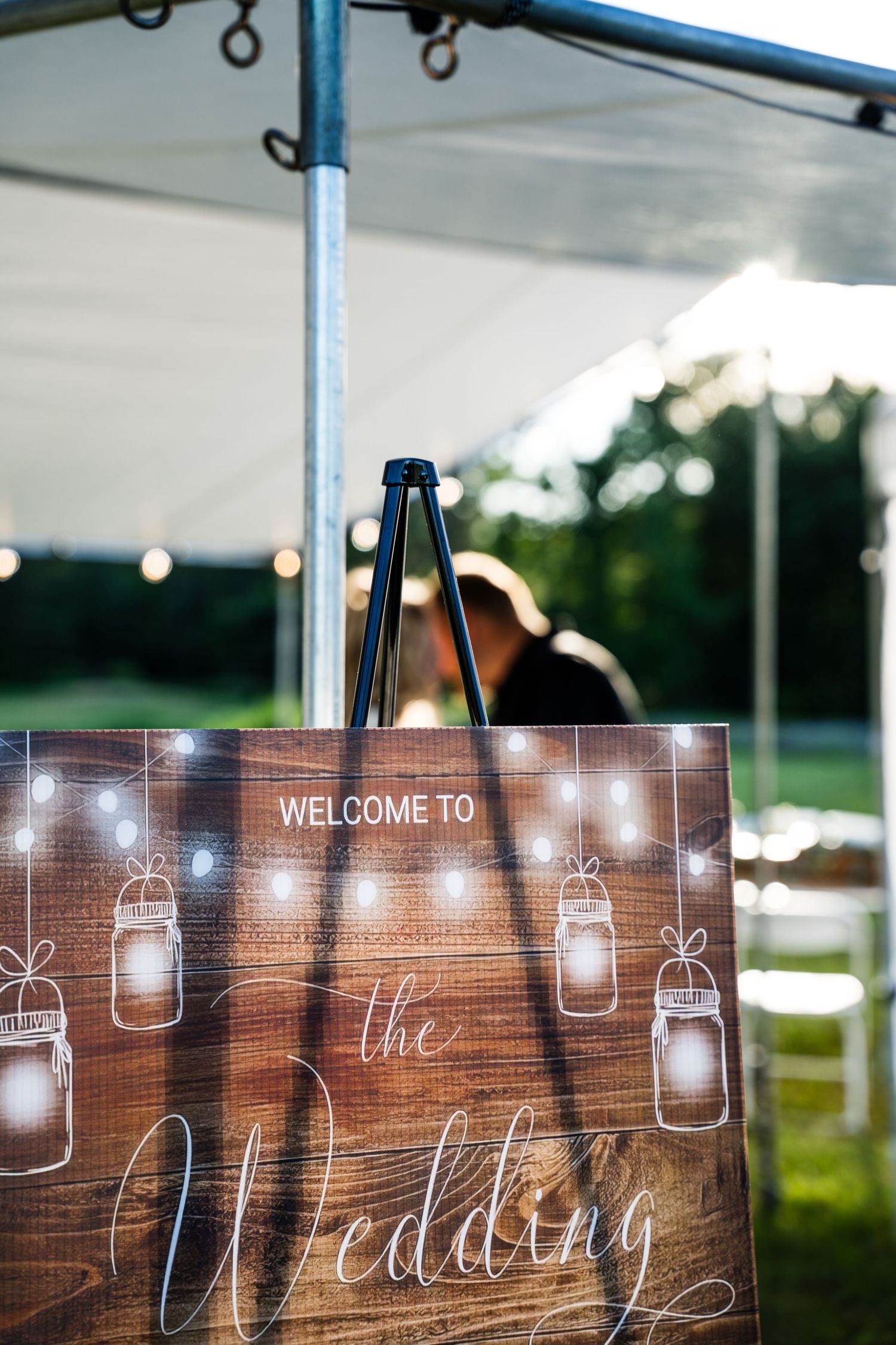Rustic wooden wedding welcome sign on easel under white tent with mason jar string lights design.