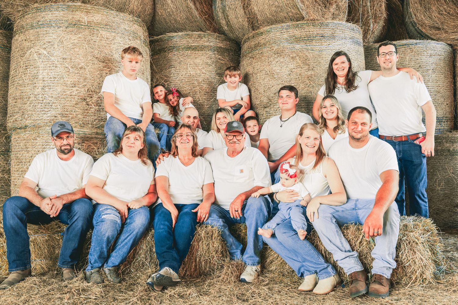 Large family in white shirts and jeans posing together in front of hay bales for a rustic outdoor portrait.