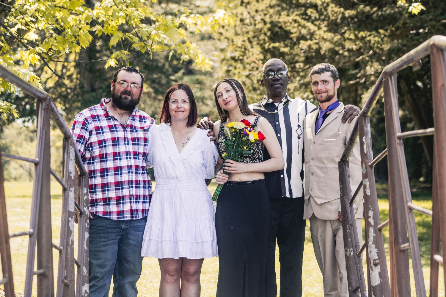 Group of five friends posing together on a bridge outdoors, with one woman holding a colorful bouquet of flowers.