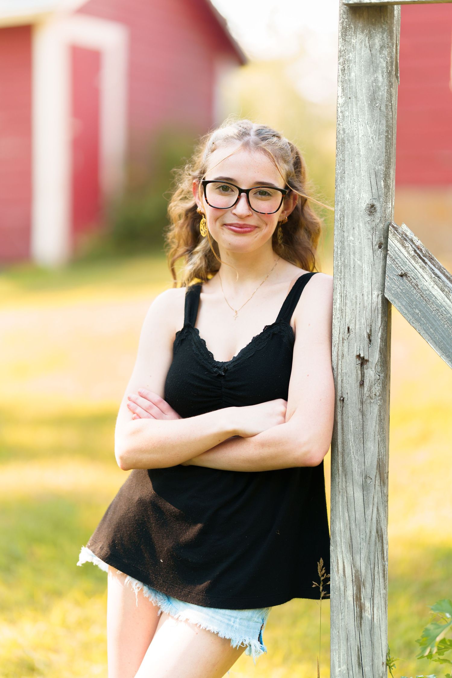 Young woman with glasses in black tank top and denim shorts leaning against wooden fence post near red barn.
