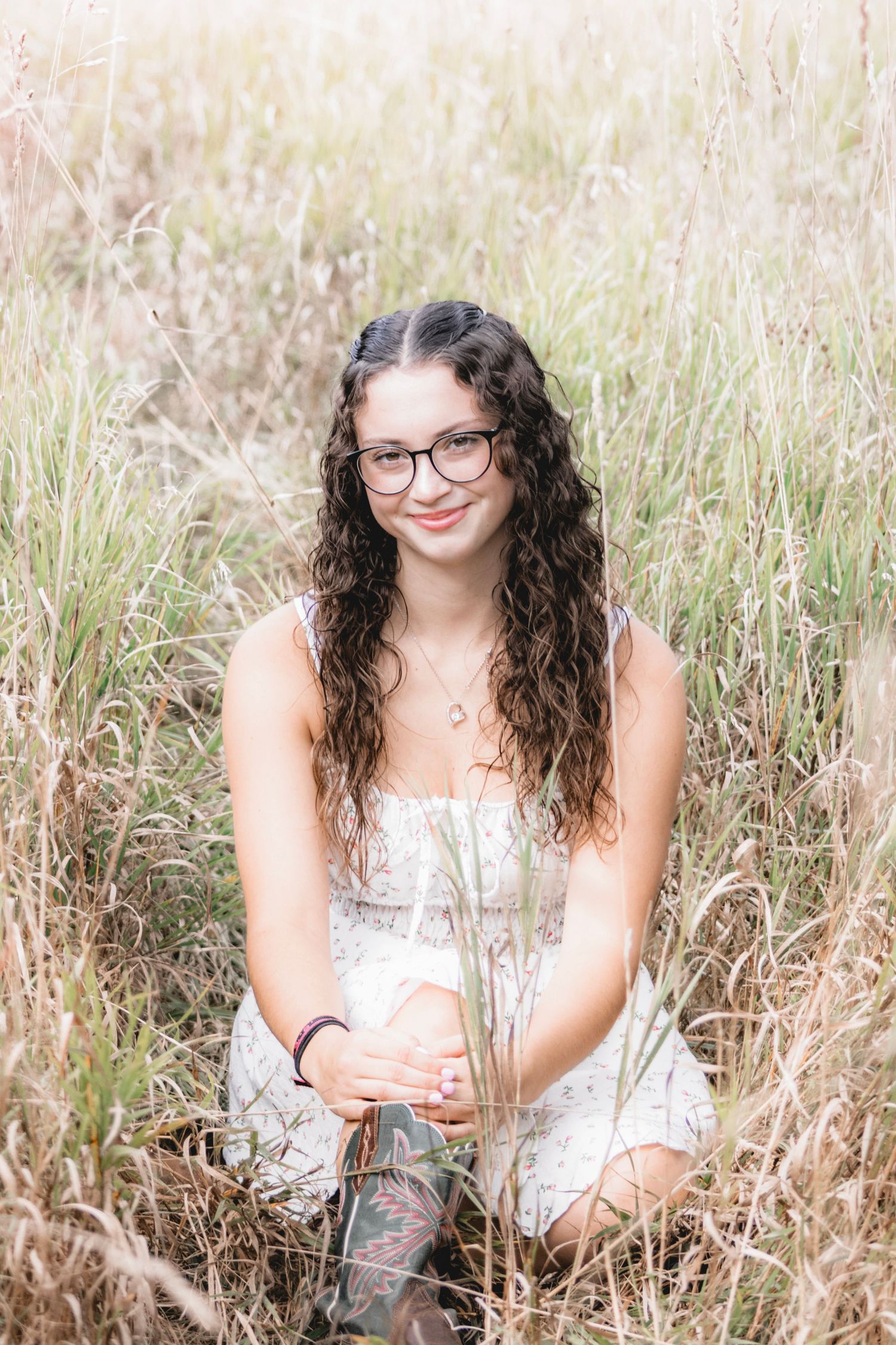 Young woman with curly hair and glasses sits smiling in tall grass wearing a white dress and boots.