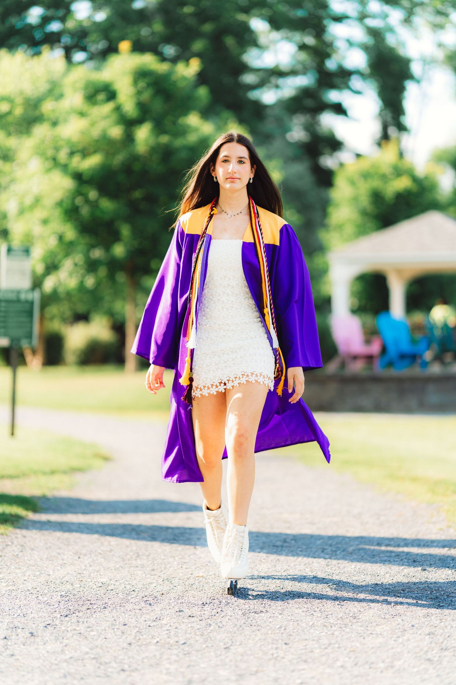 Female graduate in purple gown and white dress walking confidently on a sunny path.
