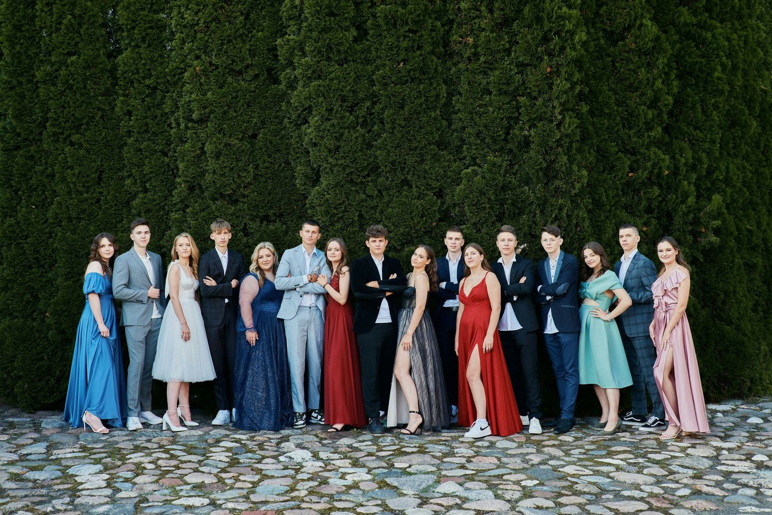 Group of teens in formal attire posing in front of a green hedge wall on a cobblestone path.
