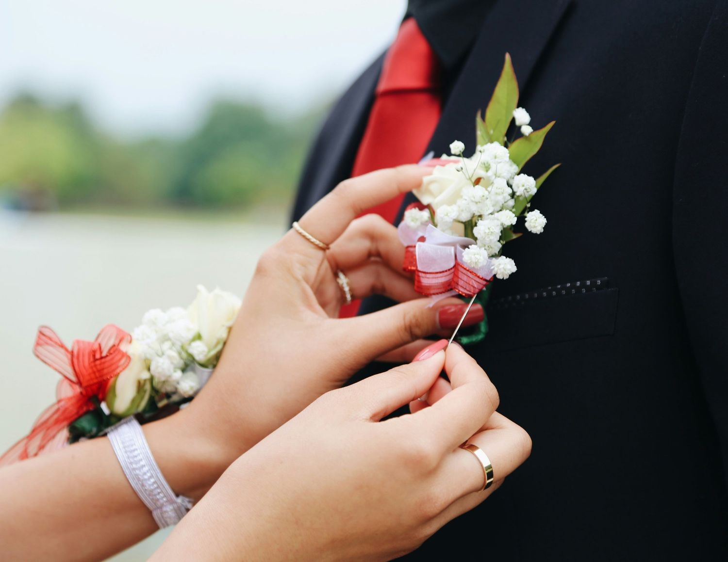 A woman with red nail polish pins a white floral boutonniere on a man's black suit jacket at prom.