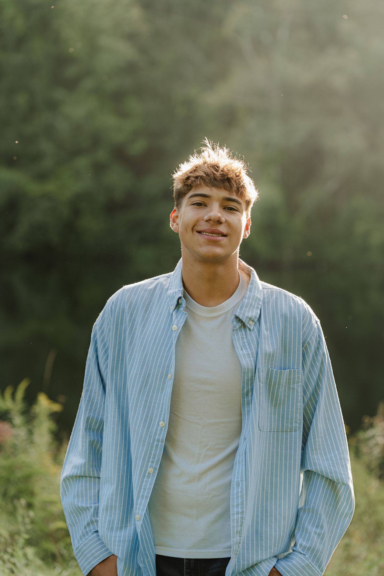 Smiling teenage boy in blue striped shirt and white tee stands outdoors with sunlit green trees in background.