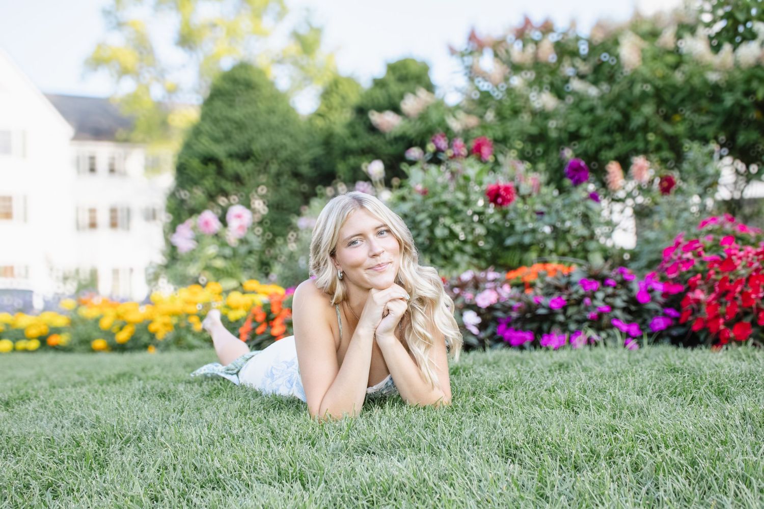 Young blonde woman lying on green grass in front of colorful flower garden on sunny day.