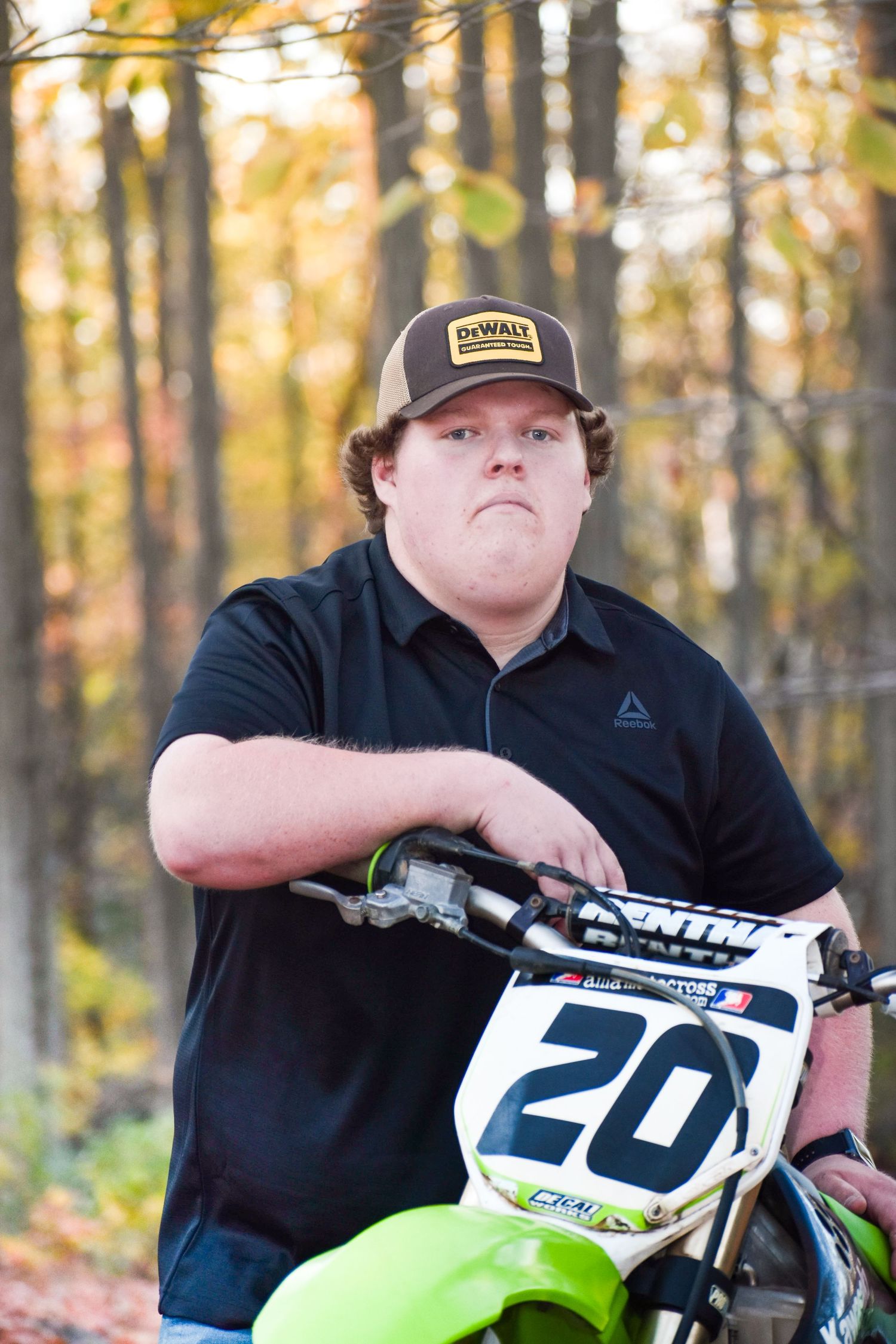 Young man in DeWalt cap and black polo leans on a green Kawasaki dirt bike numbered 20 in autumn woods.