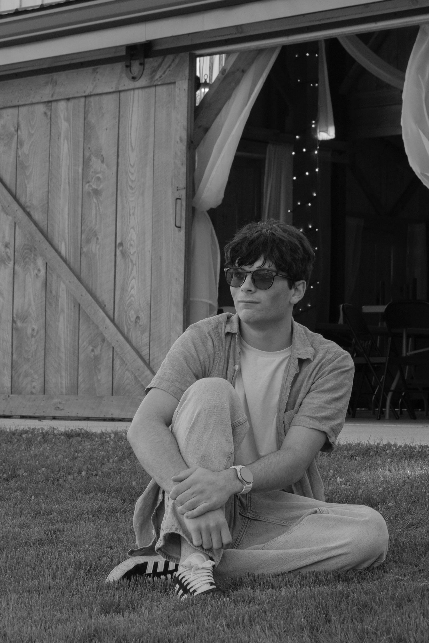 Young man in sunglasses sitting cross-legged on grass in front of a rustic wooden barn in black and white.