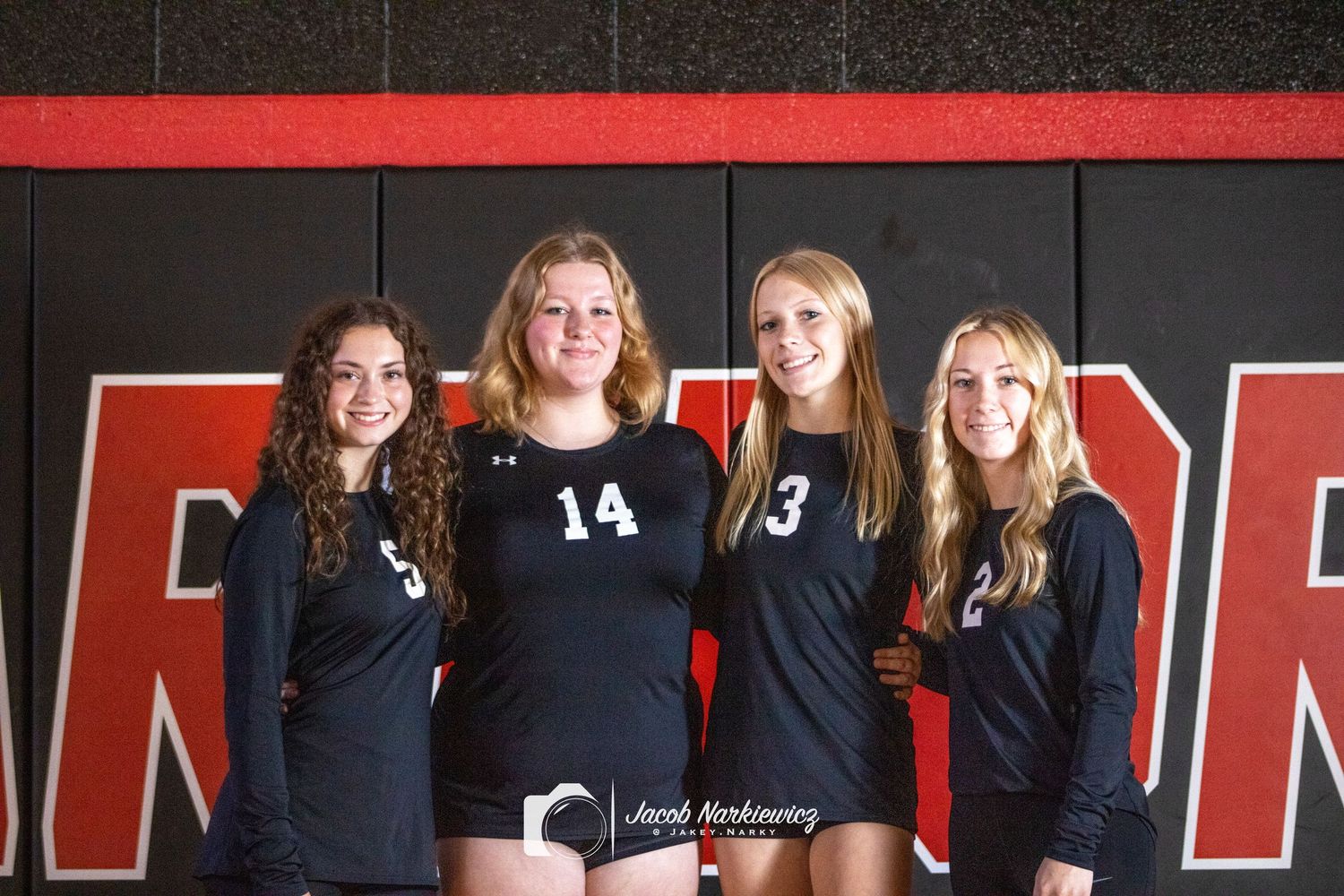 Four female volleyball players in black uniforms pose together in a gym for a team photo.