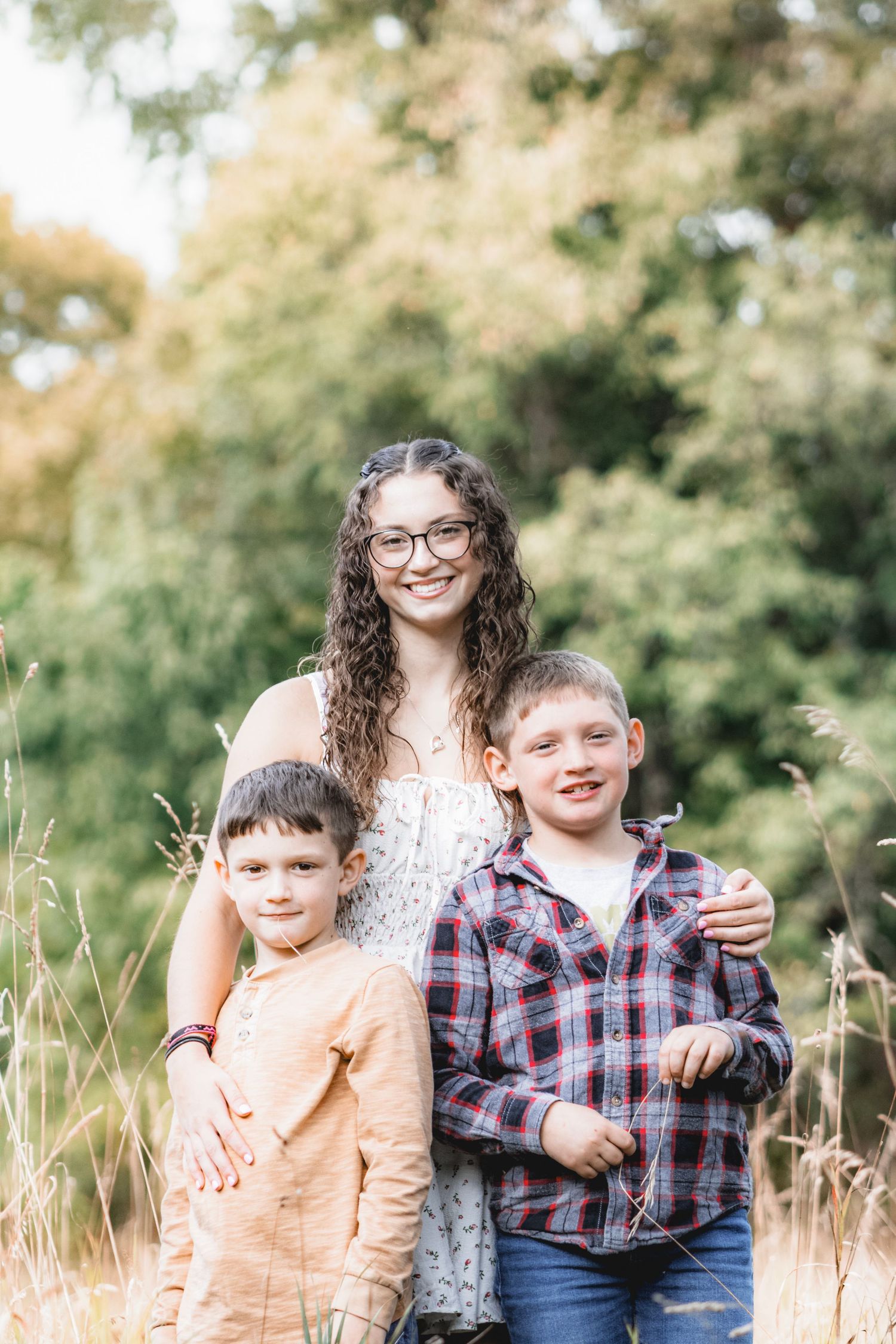A smiling teenage girl with curly hair and glasses poses outdoors with two young boys in a sunny field.