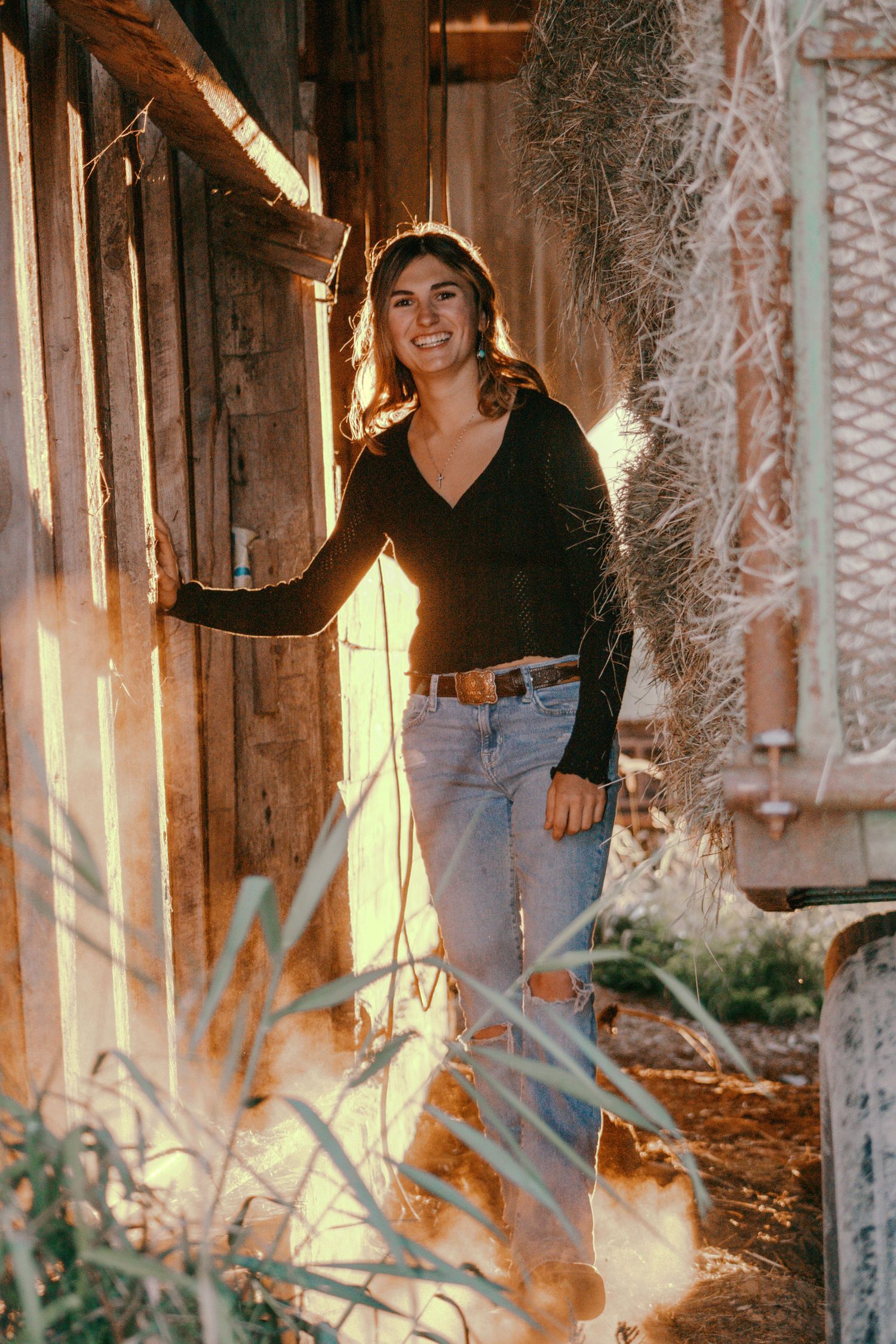 Young woman in black top and jeans smiling in golden hour sunlight near rustic wooden building.