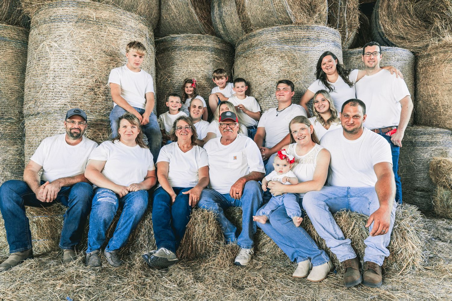 Large family group photo in front of hay bales, all wearing white shirts and jeans on a farm.
