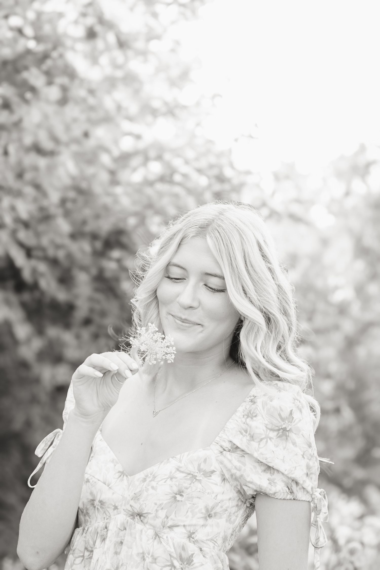 Black and white portrait of a smiling blonde woman smelling a small flower outdoors among trees.