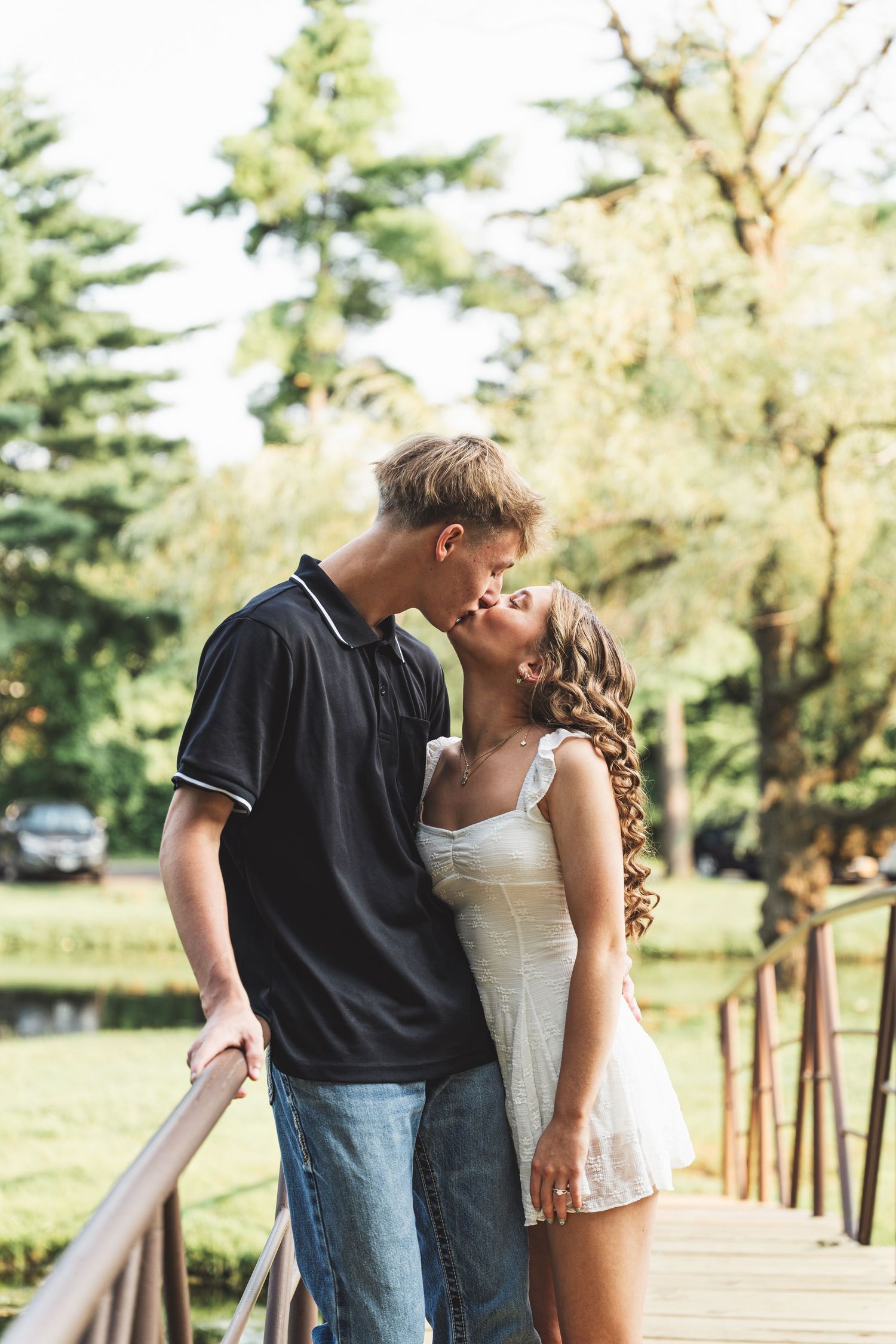 A young couple sharing a romantic kiss on a wooden bridge in a lush green park on a sunny day.