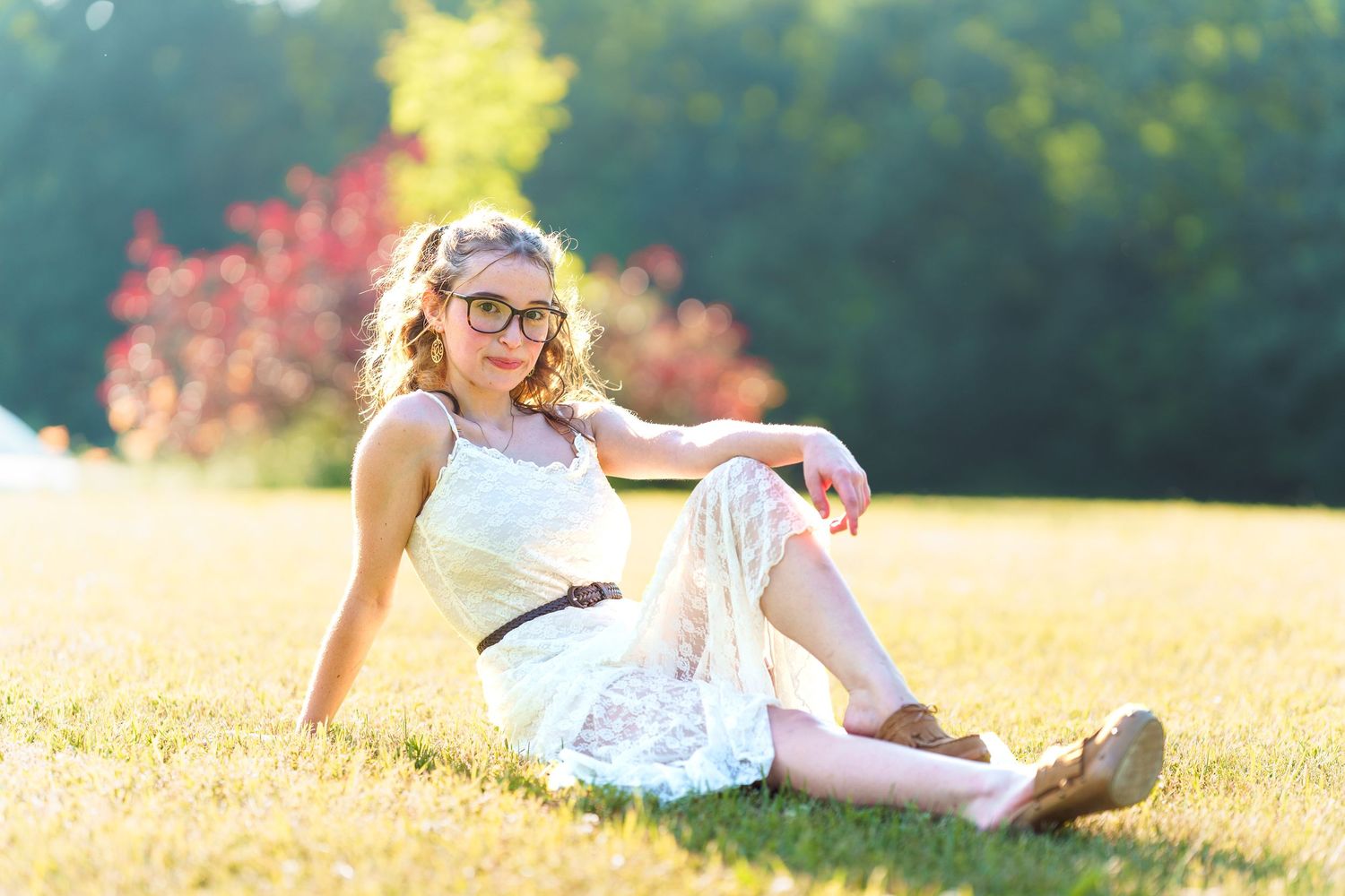 Young woman with curly hair and glasses in white lace dress sitting on green grass in sunny park.