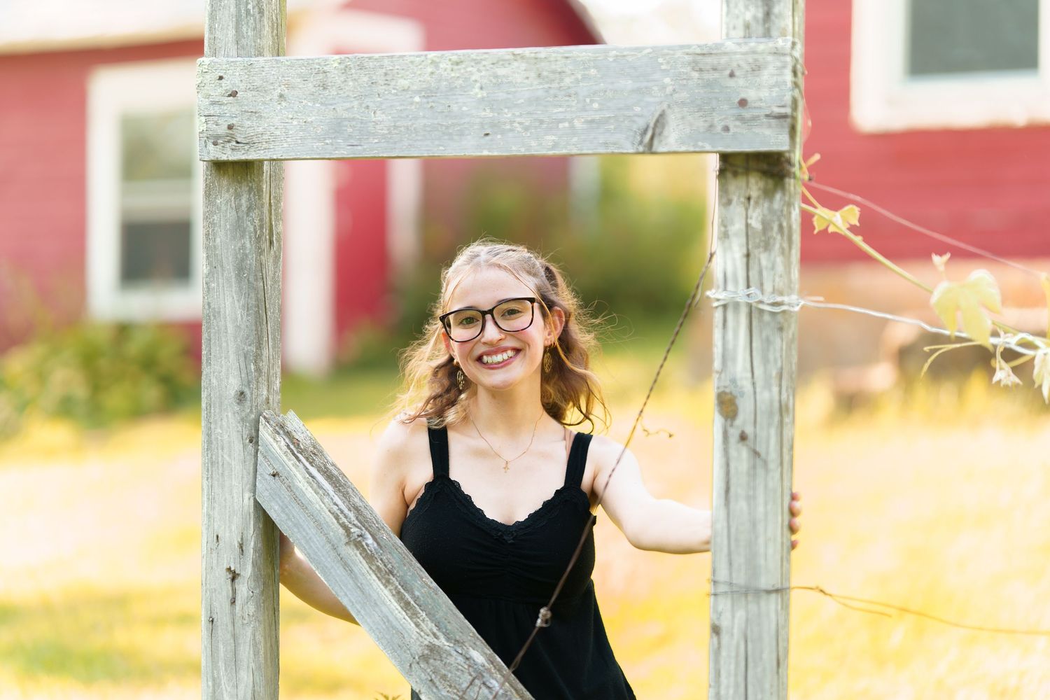 Young woman with glasses smiling outdoors near rustic wooden frame with red barn in background.